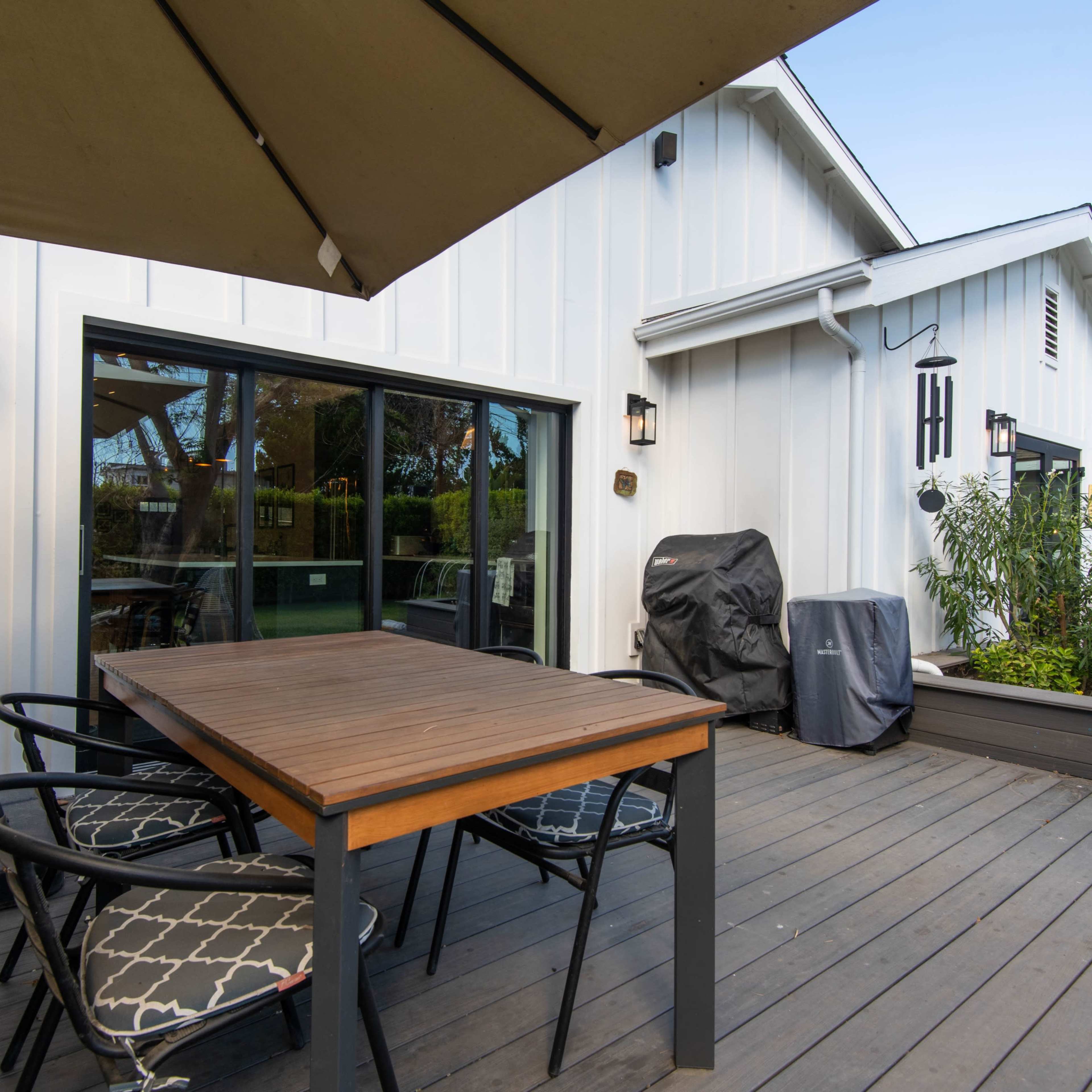 A wooden dining table with black chairs is set on a deck adjacent to a house, featuring a grill covered with a tarp.