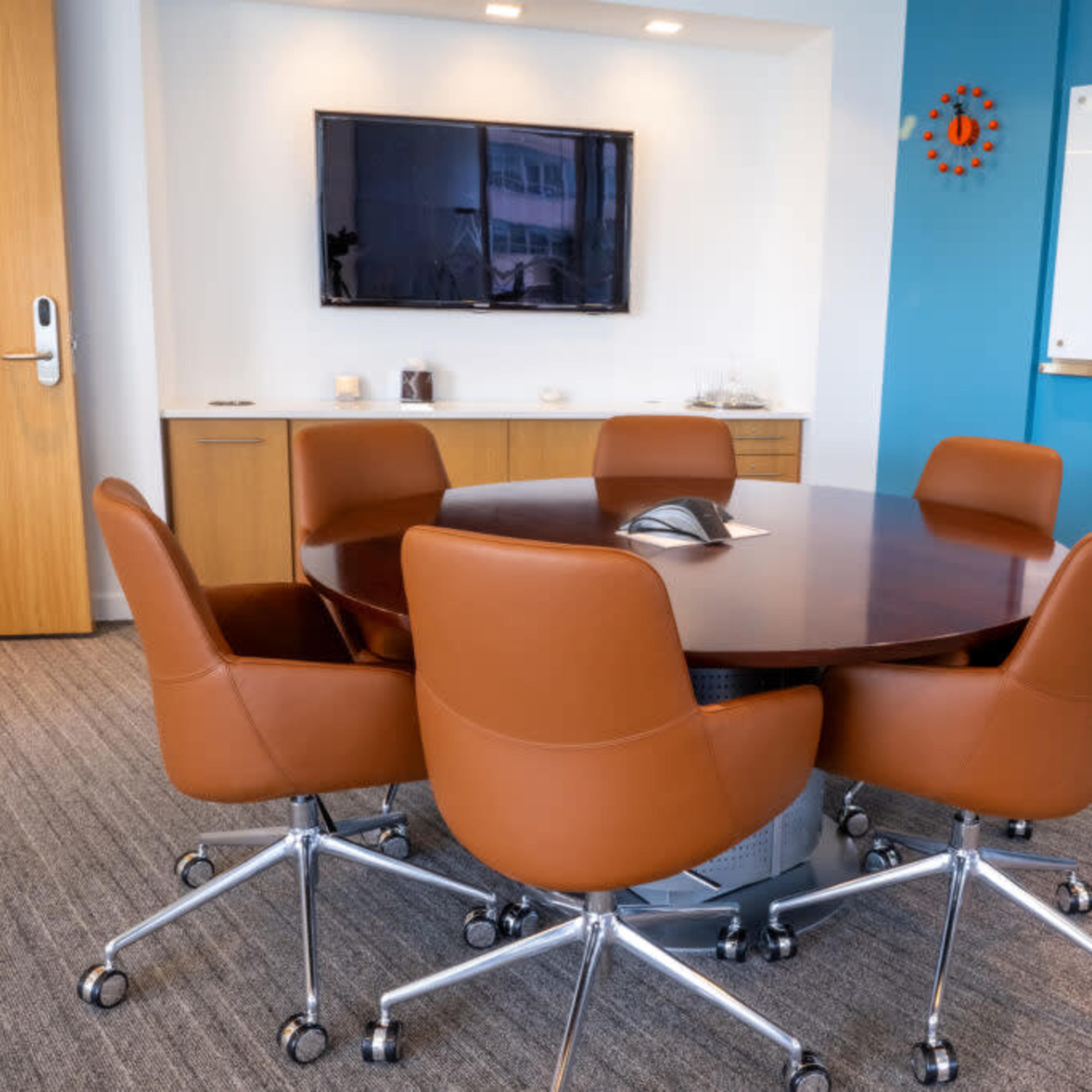 A round conference table with brown ergonomic chairs is set in a brightly lit meeting room featuring a wall-mounted TV and a whiteboard.