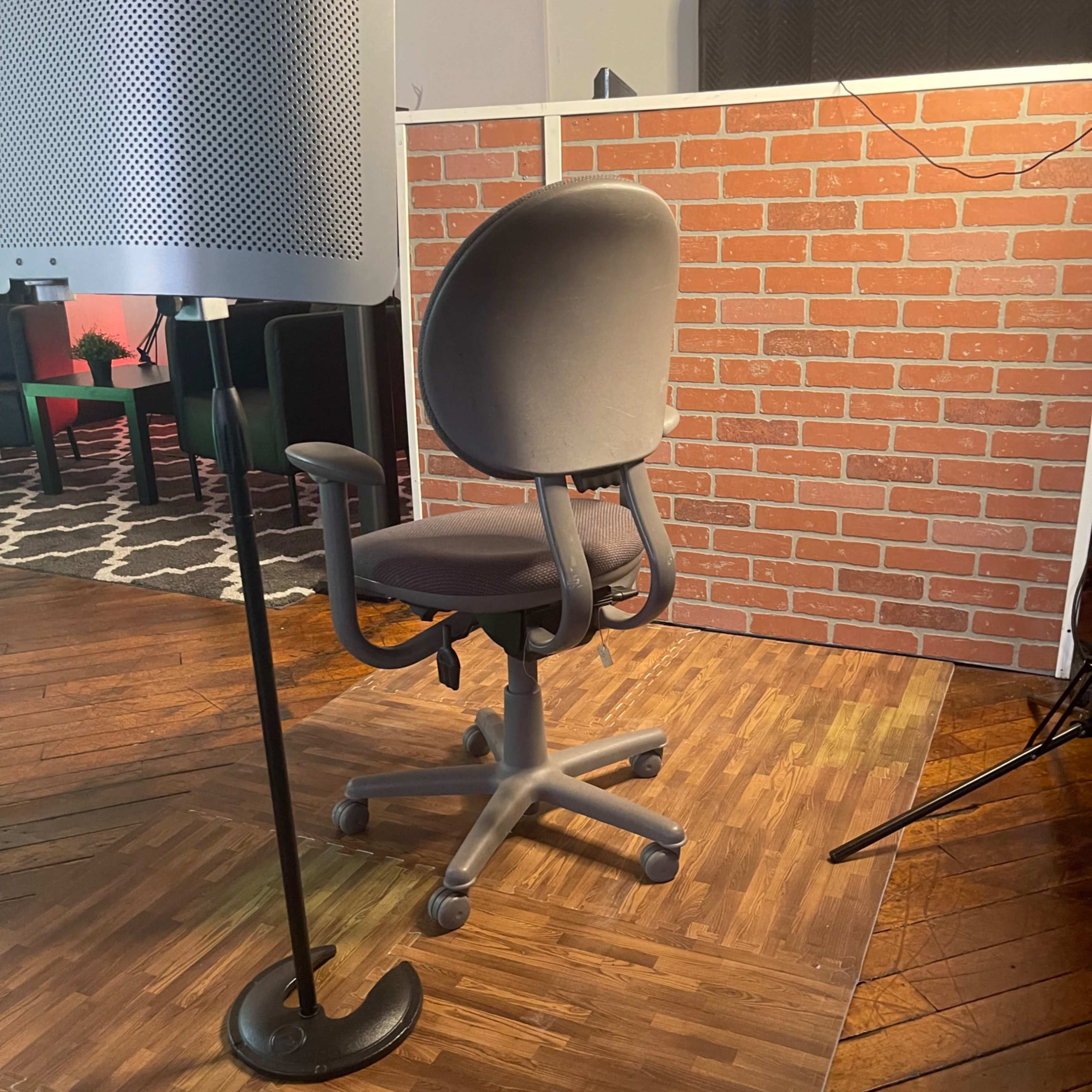A swivel office chair is positioned on a wooden floor in front of a brick-patterned backdrop.
