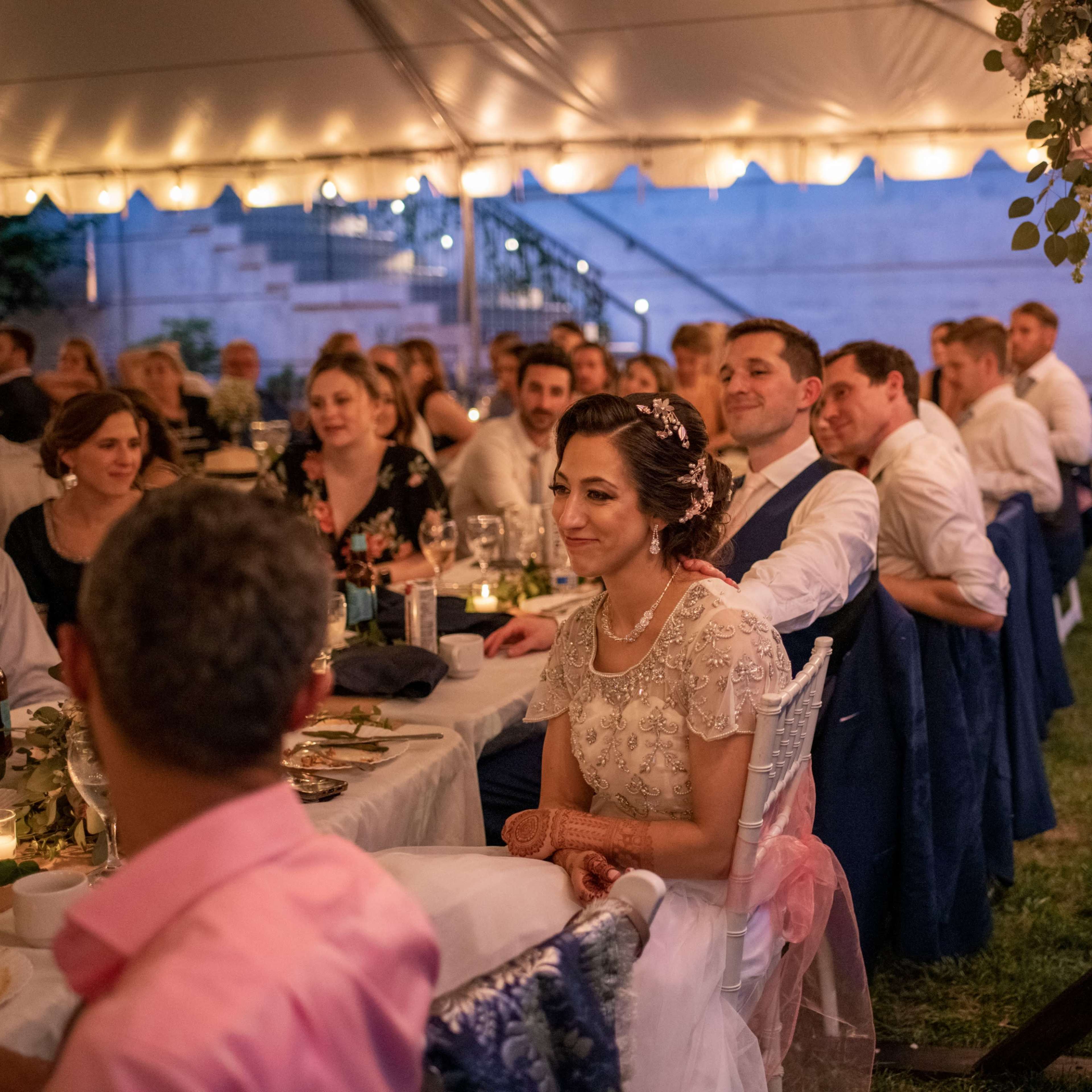 A wedding reception is taking place under a tent, where guests are seated at tables enjoying the evening with string lights overhead.