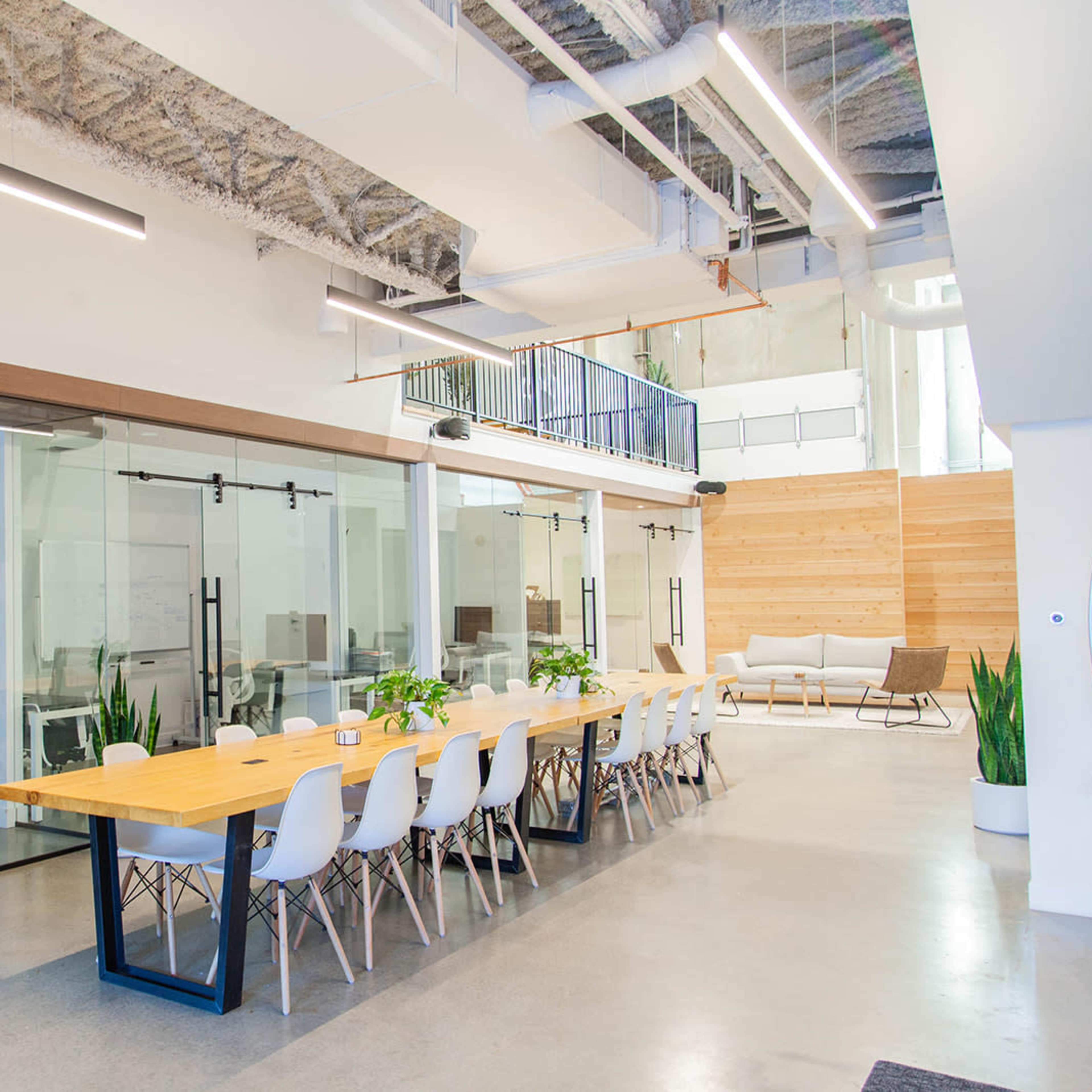 A modern office space featuring a long wooden table surrounded by white chairs, with glass-walled meeting rooms and plants lining the area.