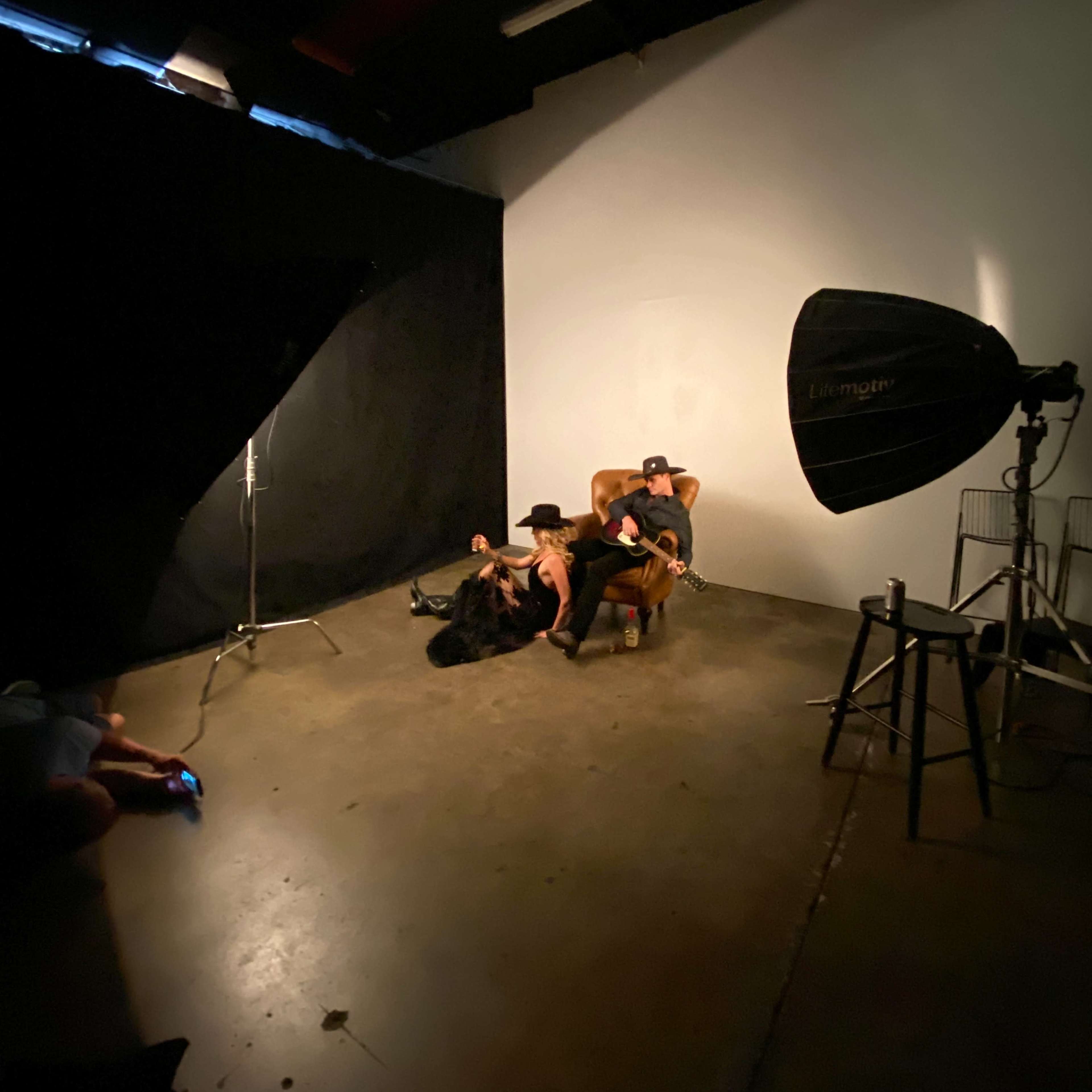 A person in a hat and a person in a chair are posed in a studio setting with a large light source and a black backdrop.
