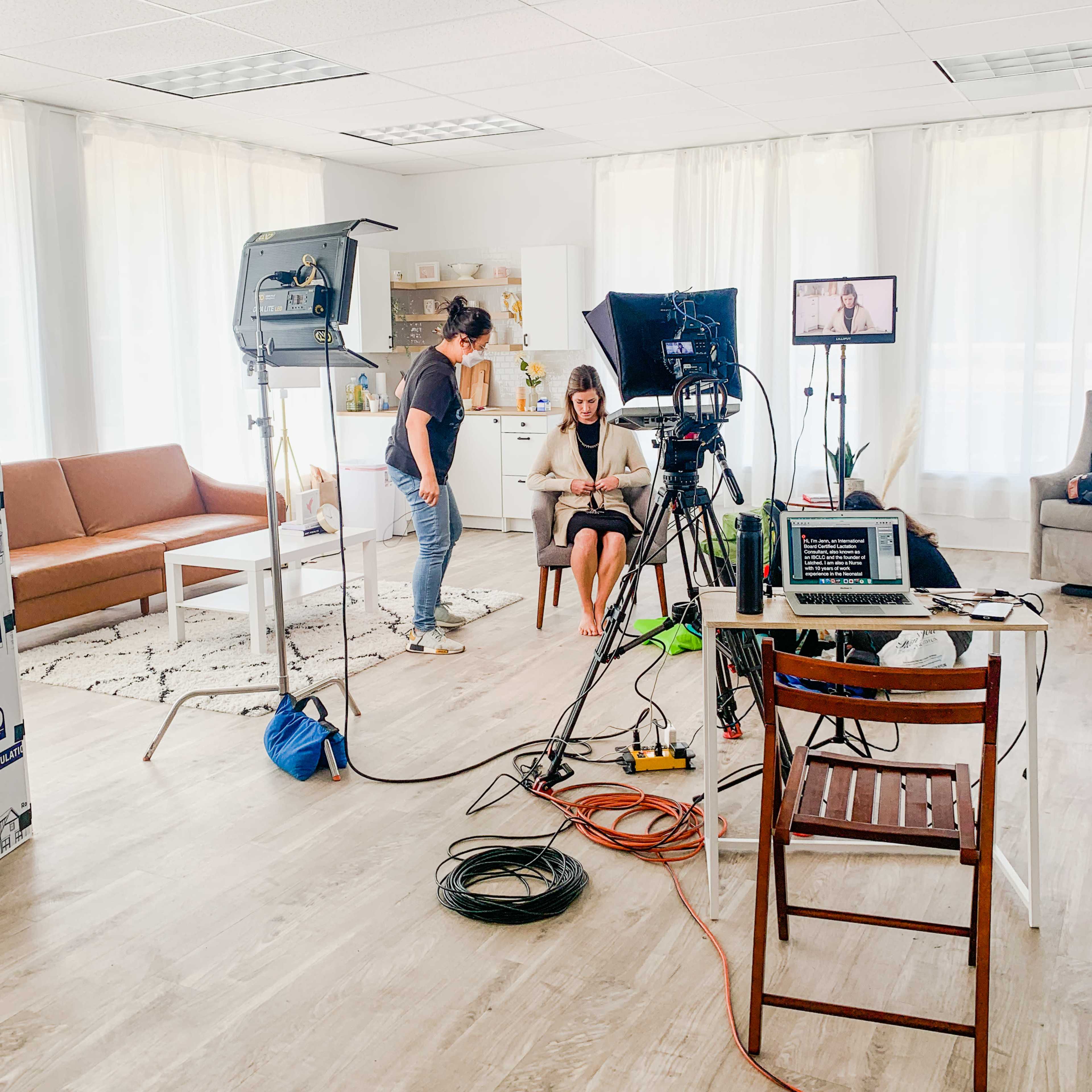 A woman sits on a chair in a bright room set up for filming, with cameras and lights arranged around her.