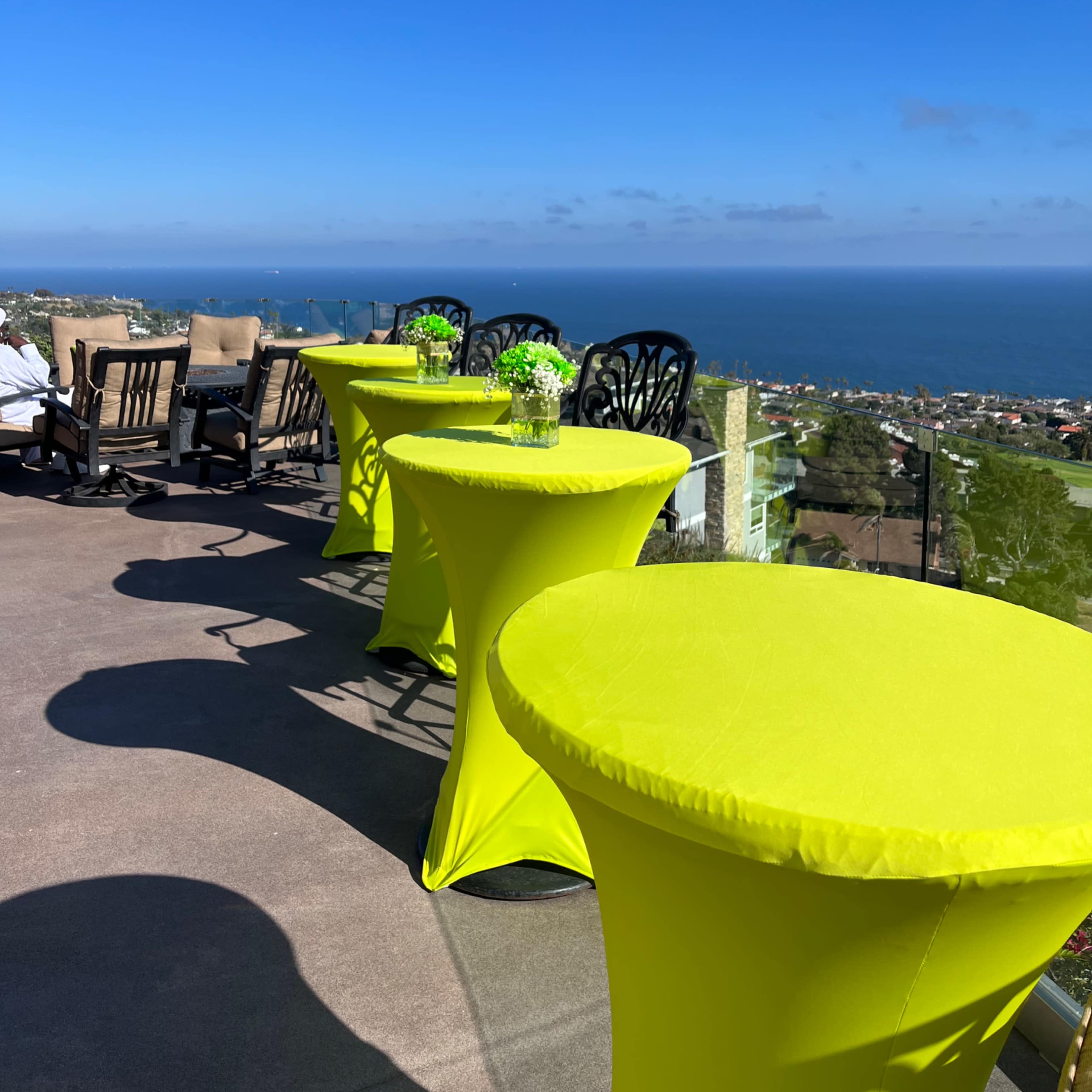 A panoramic view of the ocean is seen from a terrace with bright yellow tables and seating arrangements in the foreground.