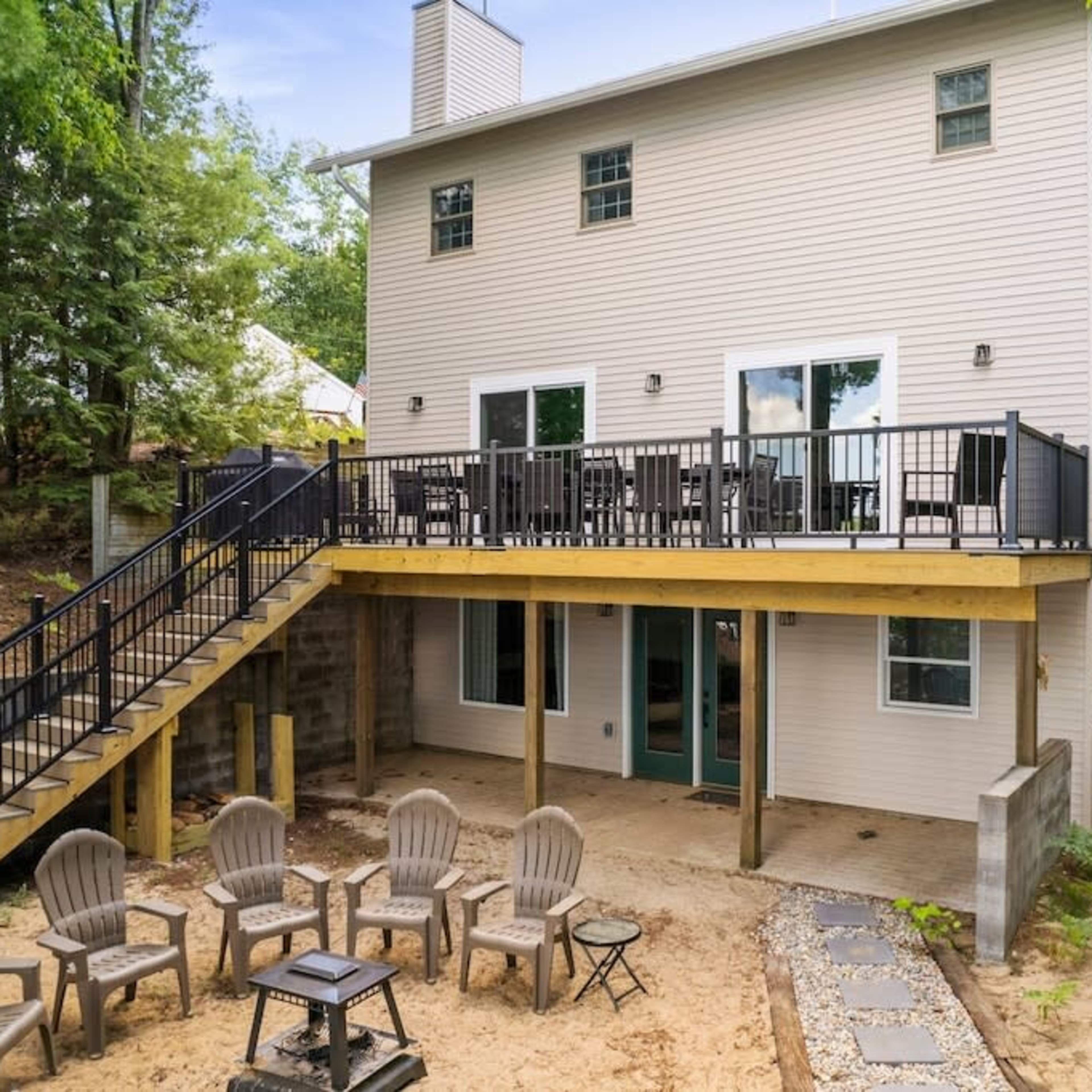 A two-story house with a spacious deck and a patio area surrounded by trees, featuring several outdoor chairs arranged in a circle.
