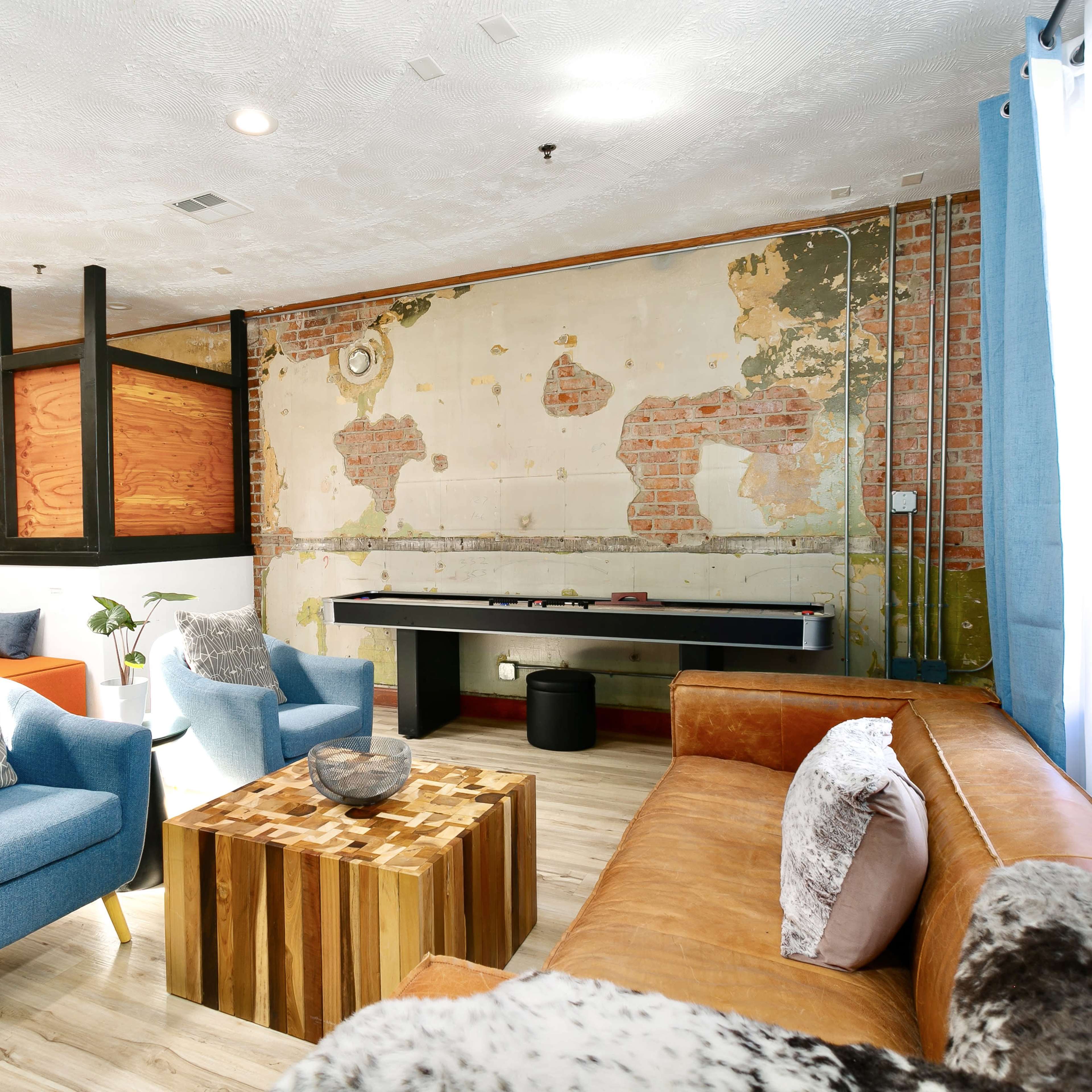 A cozy lounge area with a leather sofa, blue chairs, a wooden coffee table, and a partially exposed brick wall featuring a vintage world map.
