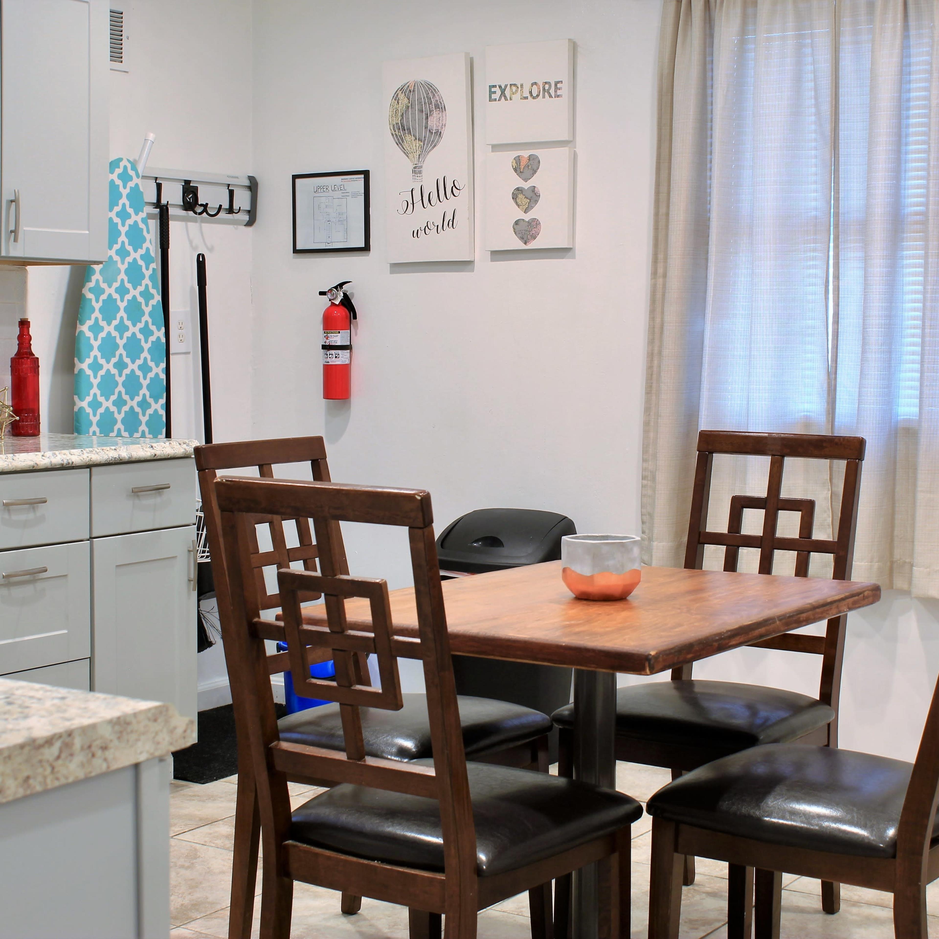 A dining area features a wooden table with four chairs, surrounded by light-colored cabinets and decorations on the walls.