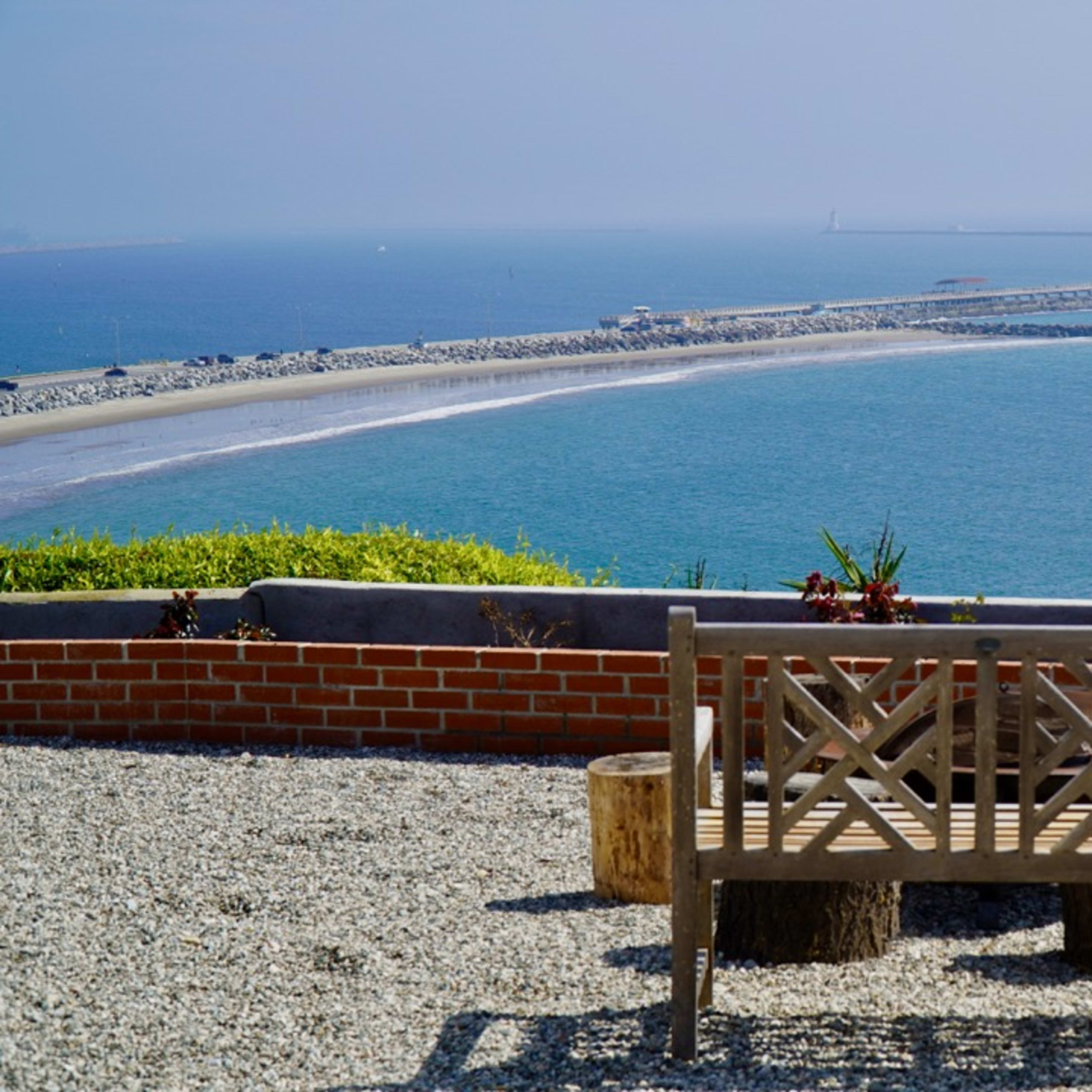 A wooden bench overlooks a rocky jetty extending into a calm blue sea under a clear sky.
