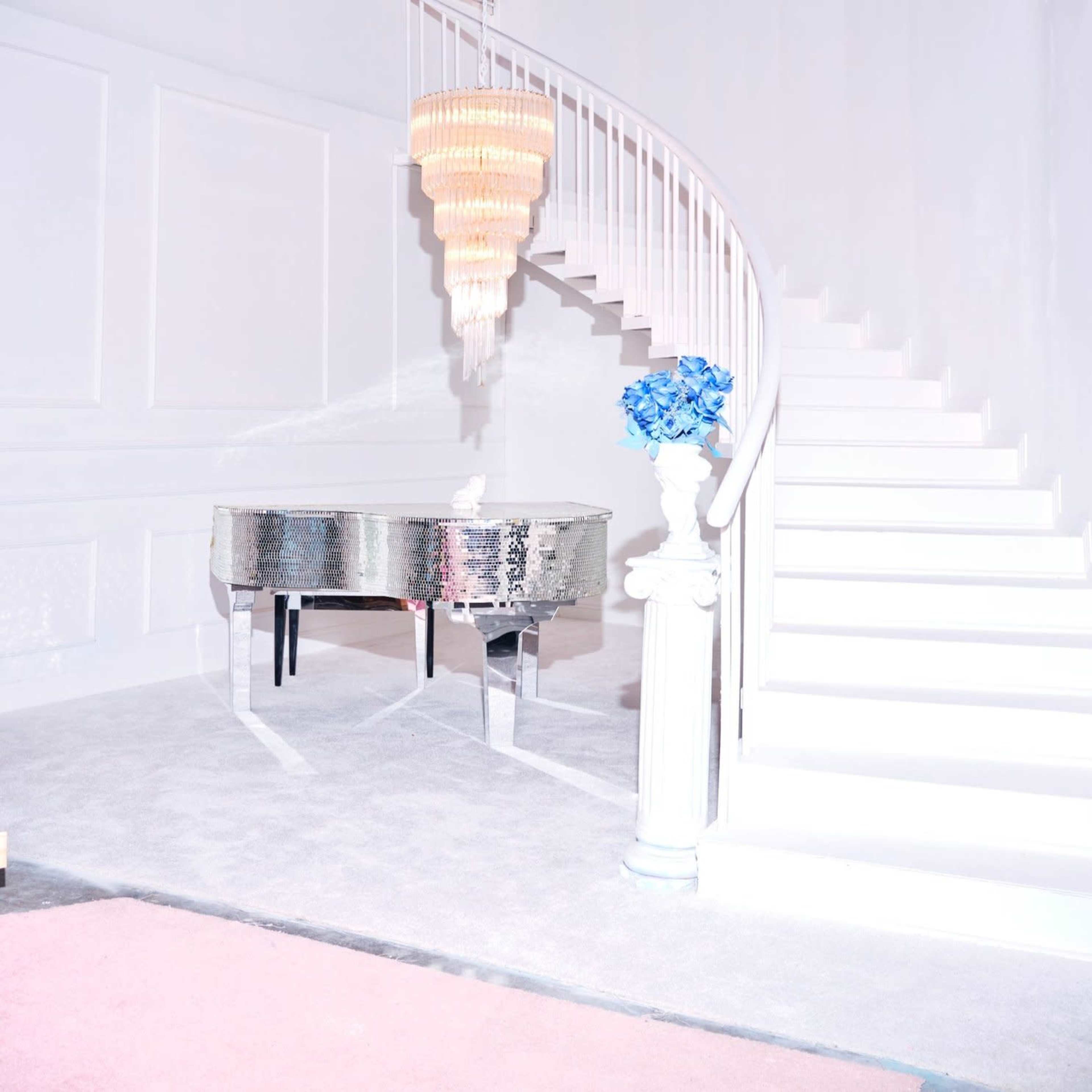 A silver grand piano sits on a plush carpet next to a blue flower arrangement, with a white staircase and a chandelier above.