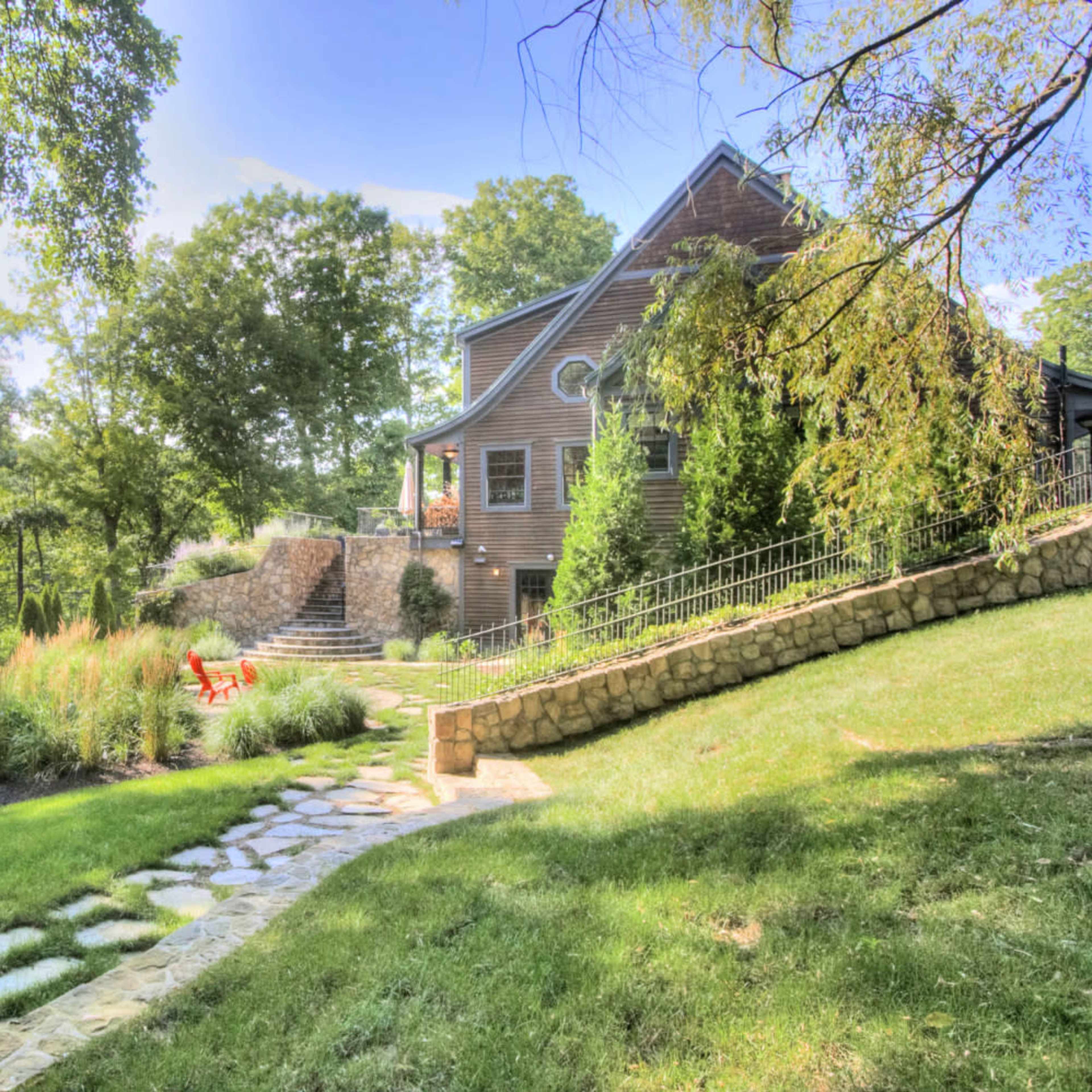 A stone pathway leads to a wooden house situated among trees and landscaping, with a patio and a red chair visible in the yard.