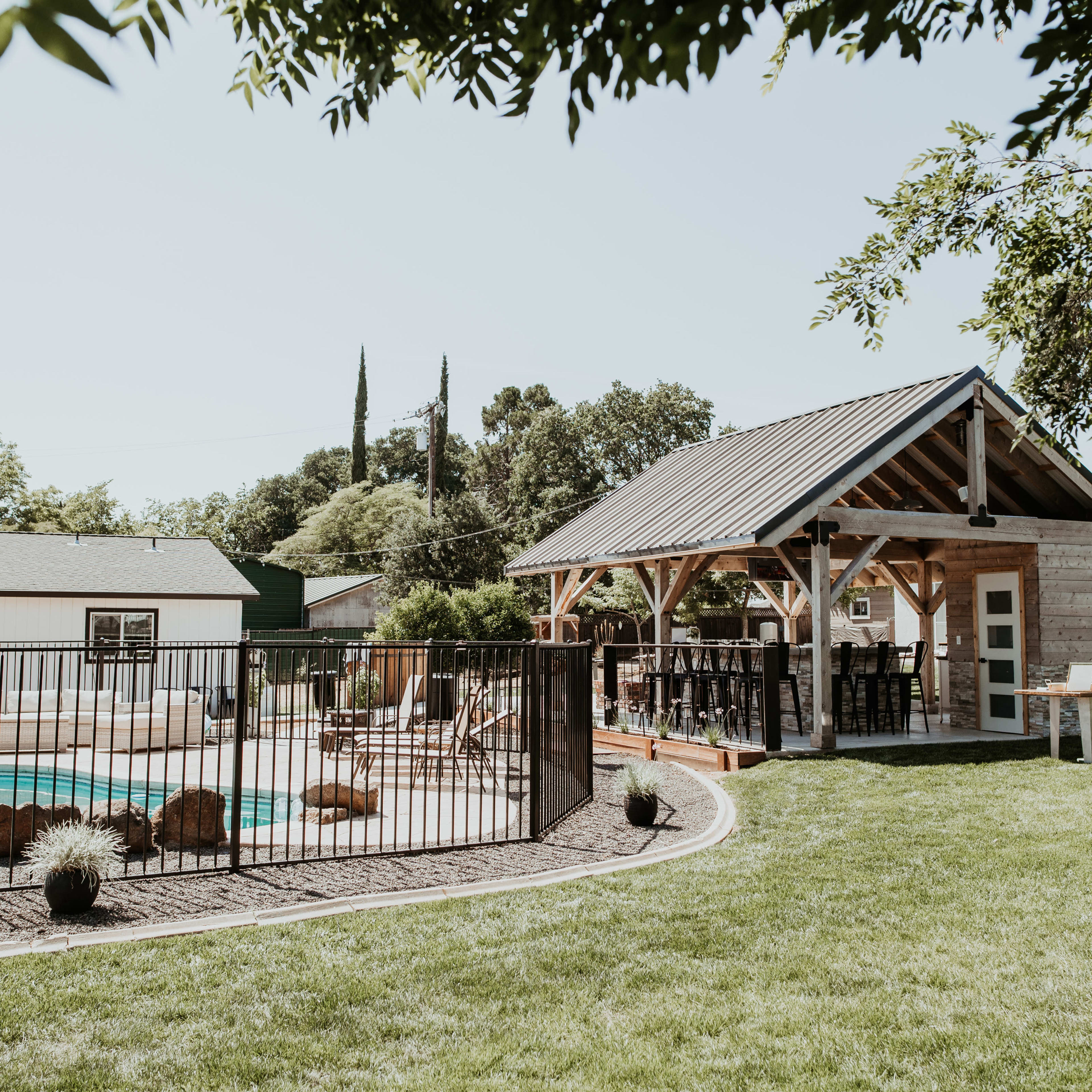 A backyard features a swimming pool surrounded by a black fence and a gazebo with a shaded seating area.