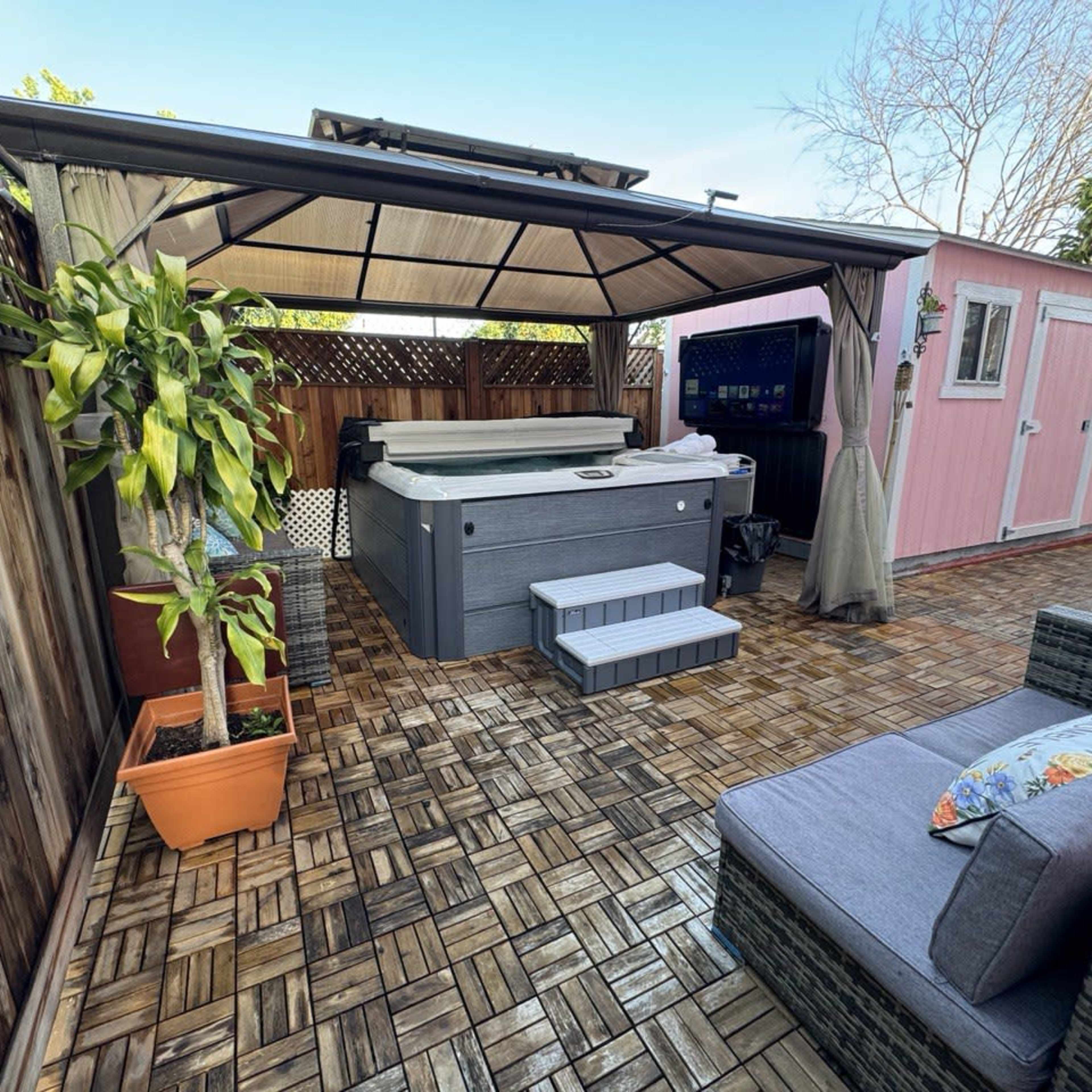 A backyard with a hot tub under a canopy, surrounded by wooden fencing, potted plants, and a pink storage shed.