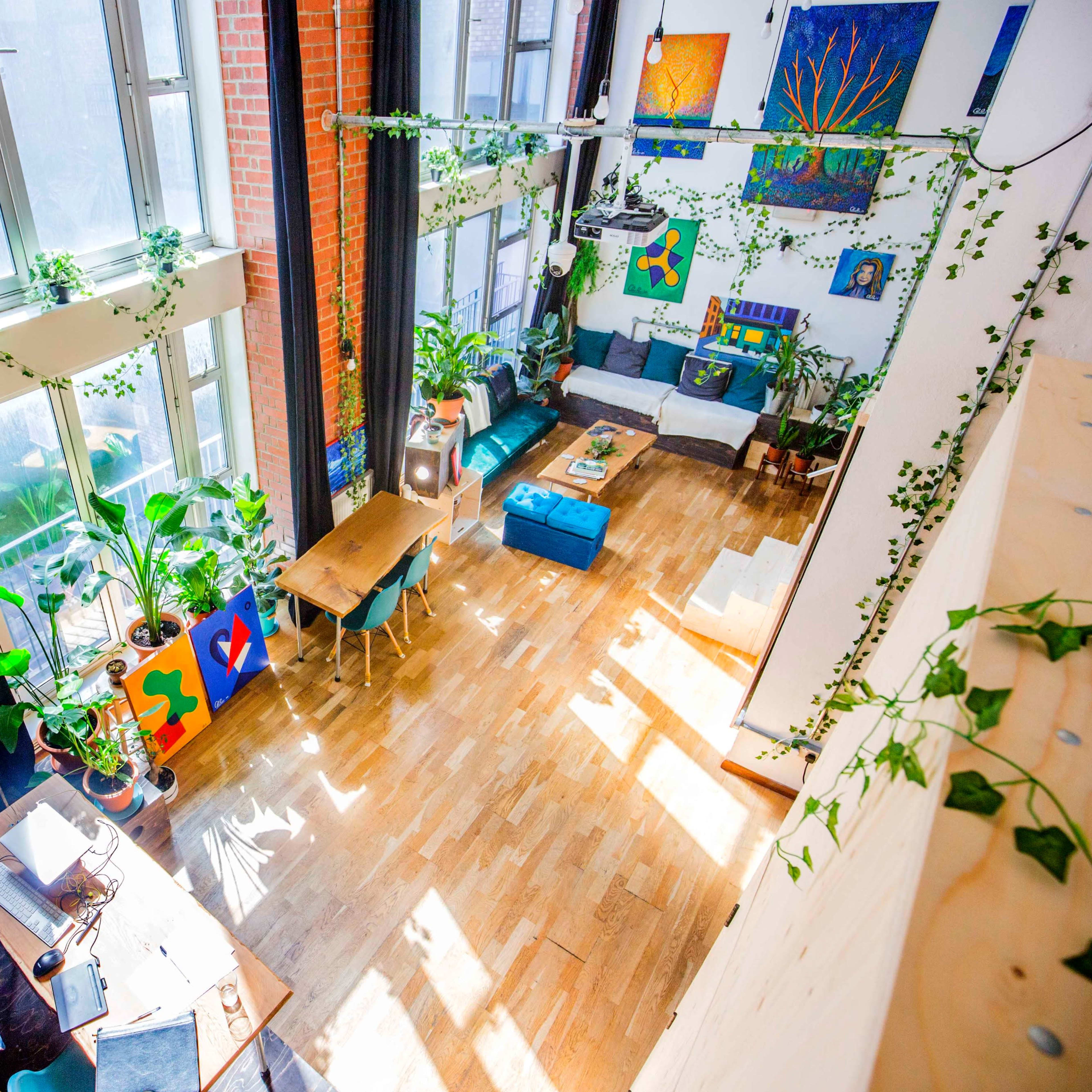 A bright and spacious room with large windows, wooden floors, plants, and colorful artwork on the walls.