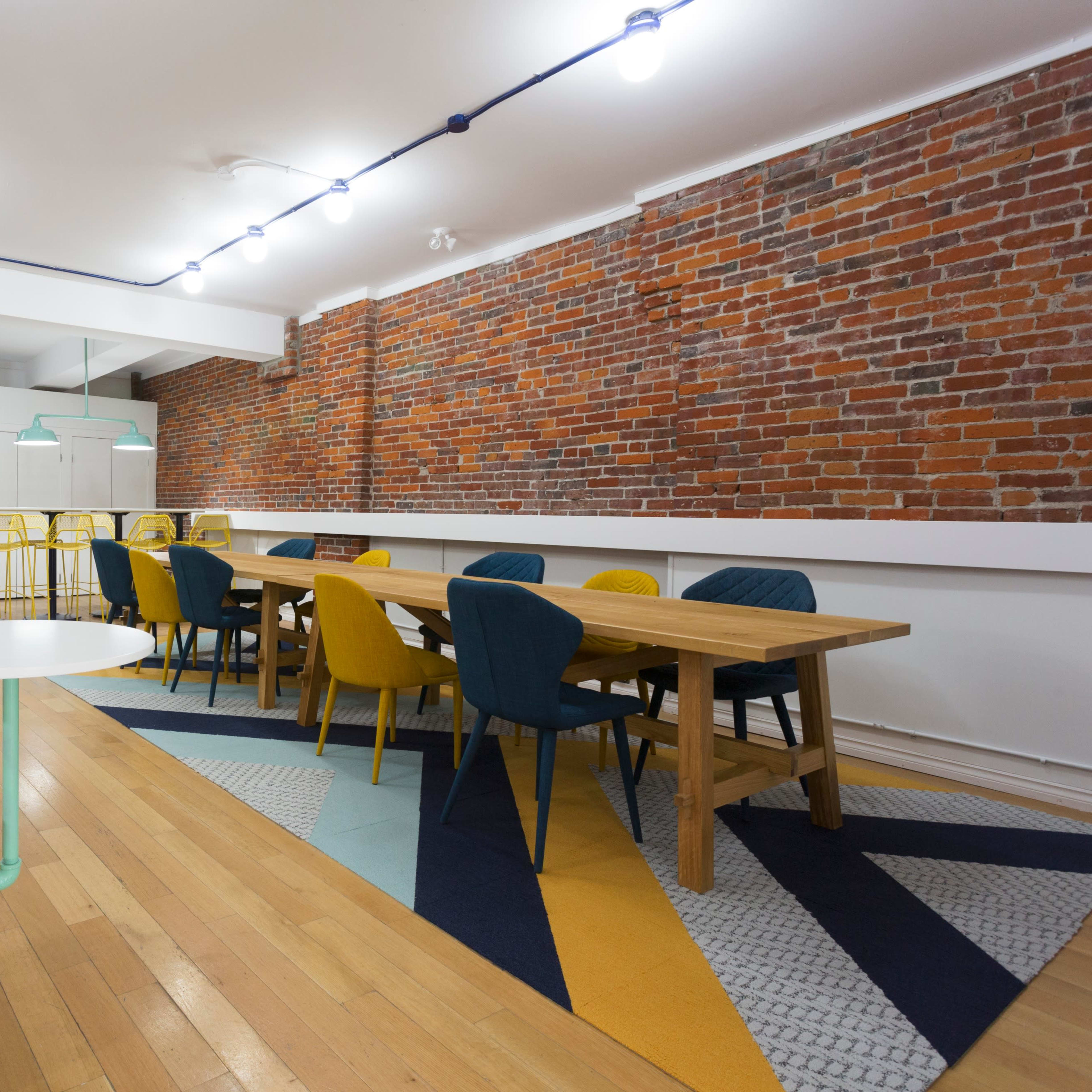 A modern, open interior space with a wooden dining table surrounded by colorful chairs, exposed brick walls, and a mix of patterned rugs on the floor.
