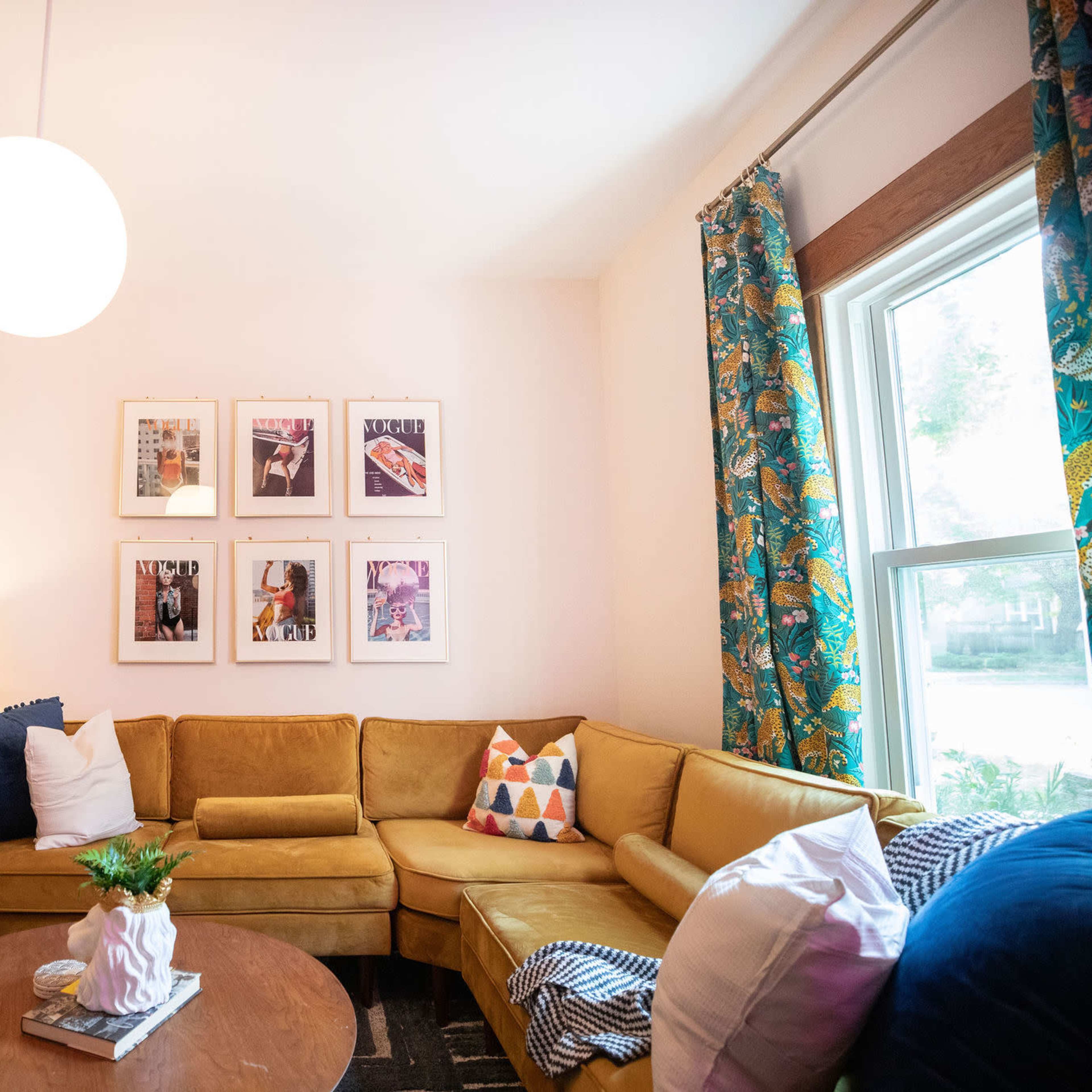 A cozy living room features a yellow sectional sofa, a round coffee table, decorative pillows, and framed artwork on the wall, with a floor lamp and a potted plant in the corner.