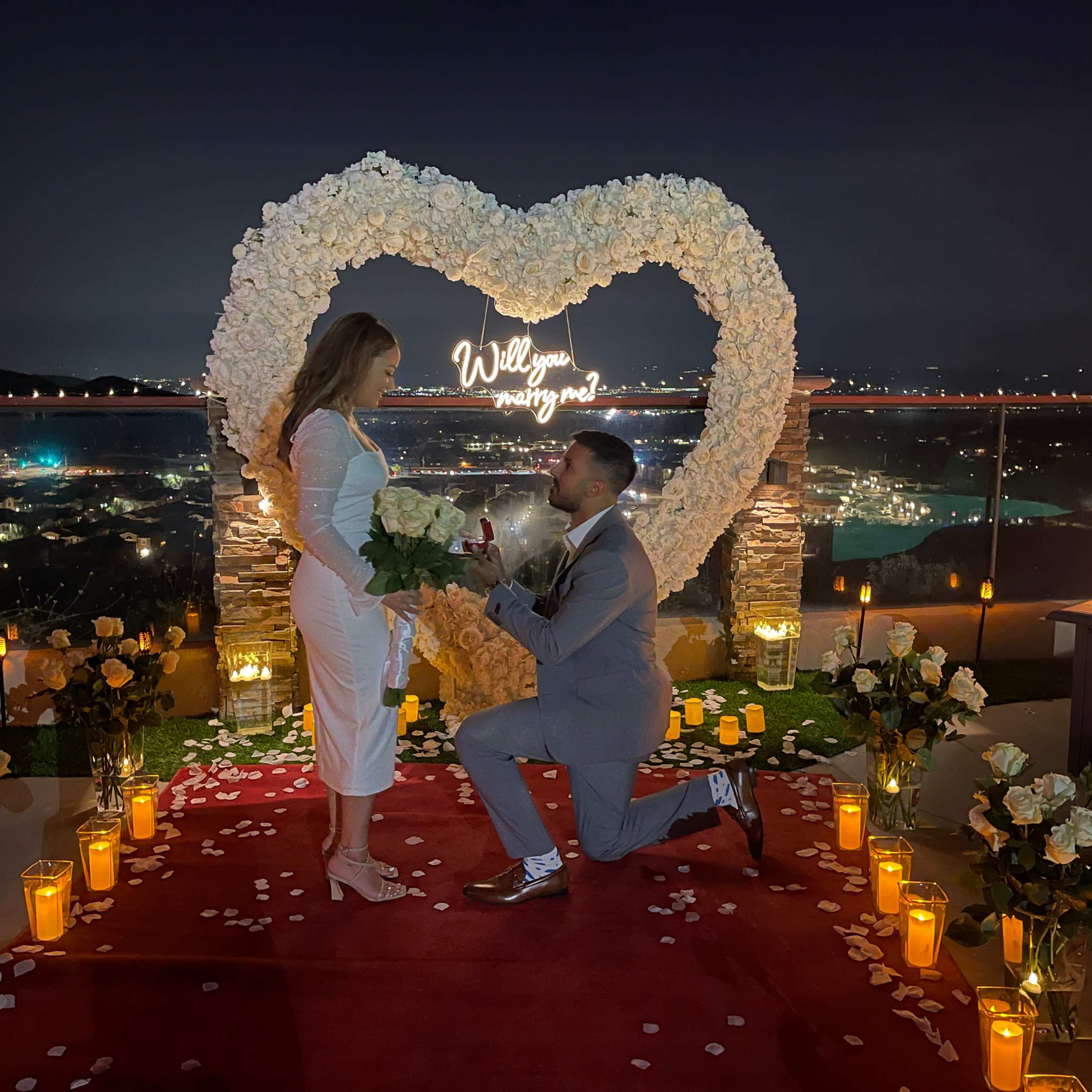 A man is proposing to a woman in front of a heart-shaped floral display, surrounded by candles and a city skyline at night.