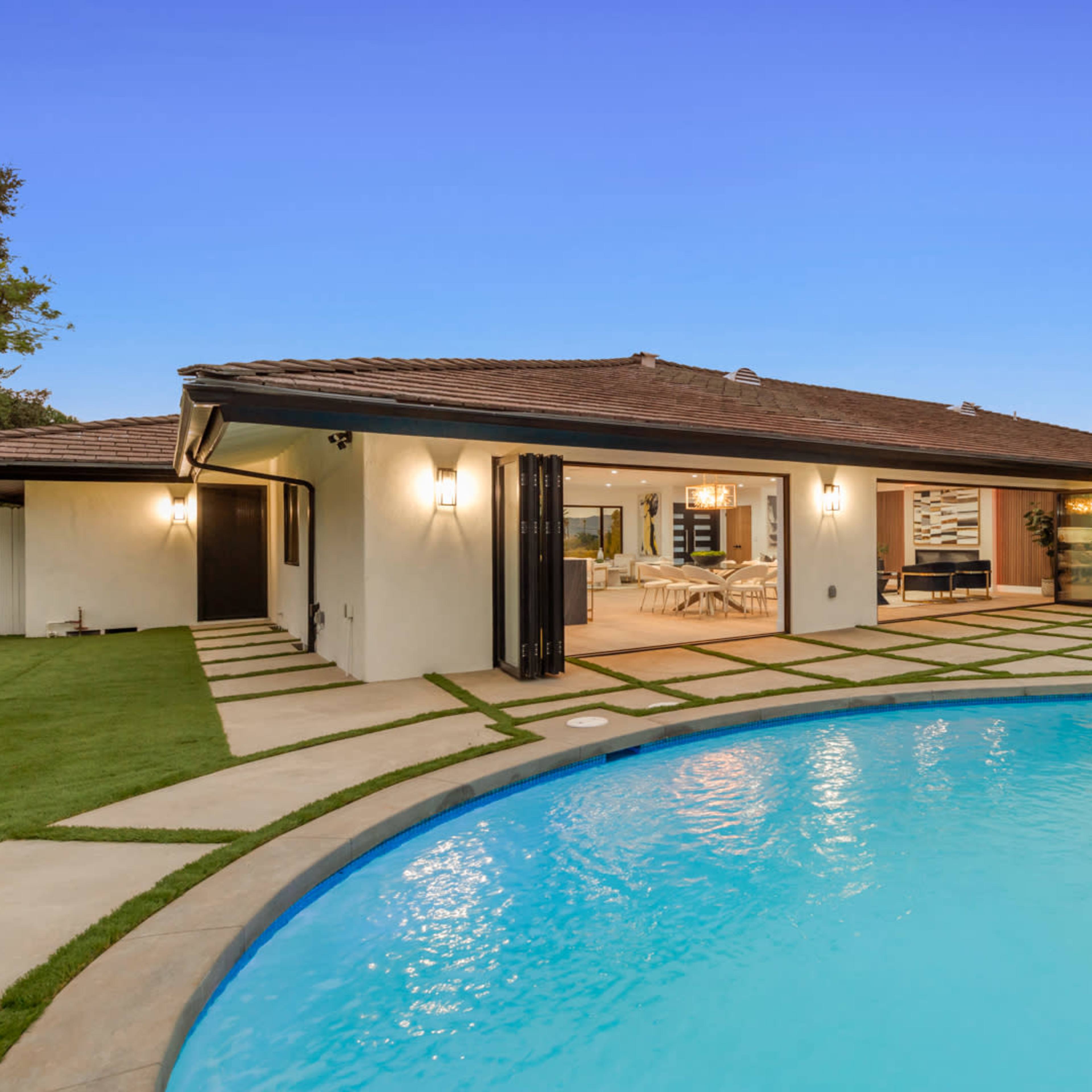 A single-story house with a tiled roof is situated next to a curved swimming pool, featuring a stone patio and landscaped green surroundings.