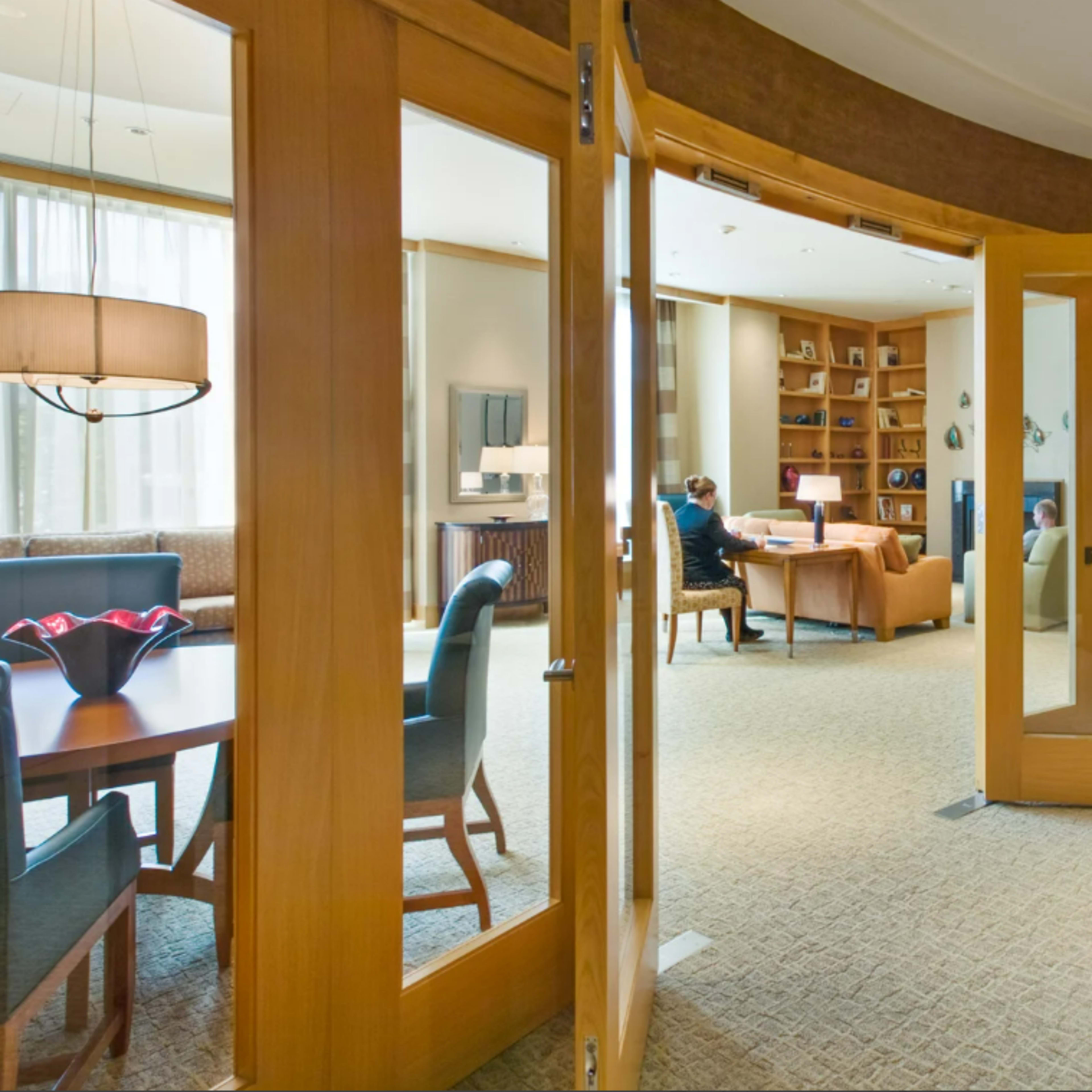 A spacious lounge area features wooden doors leading into a well-lit room with a seating area and a person working at a desk.