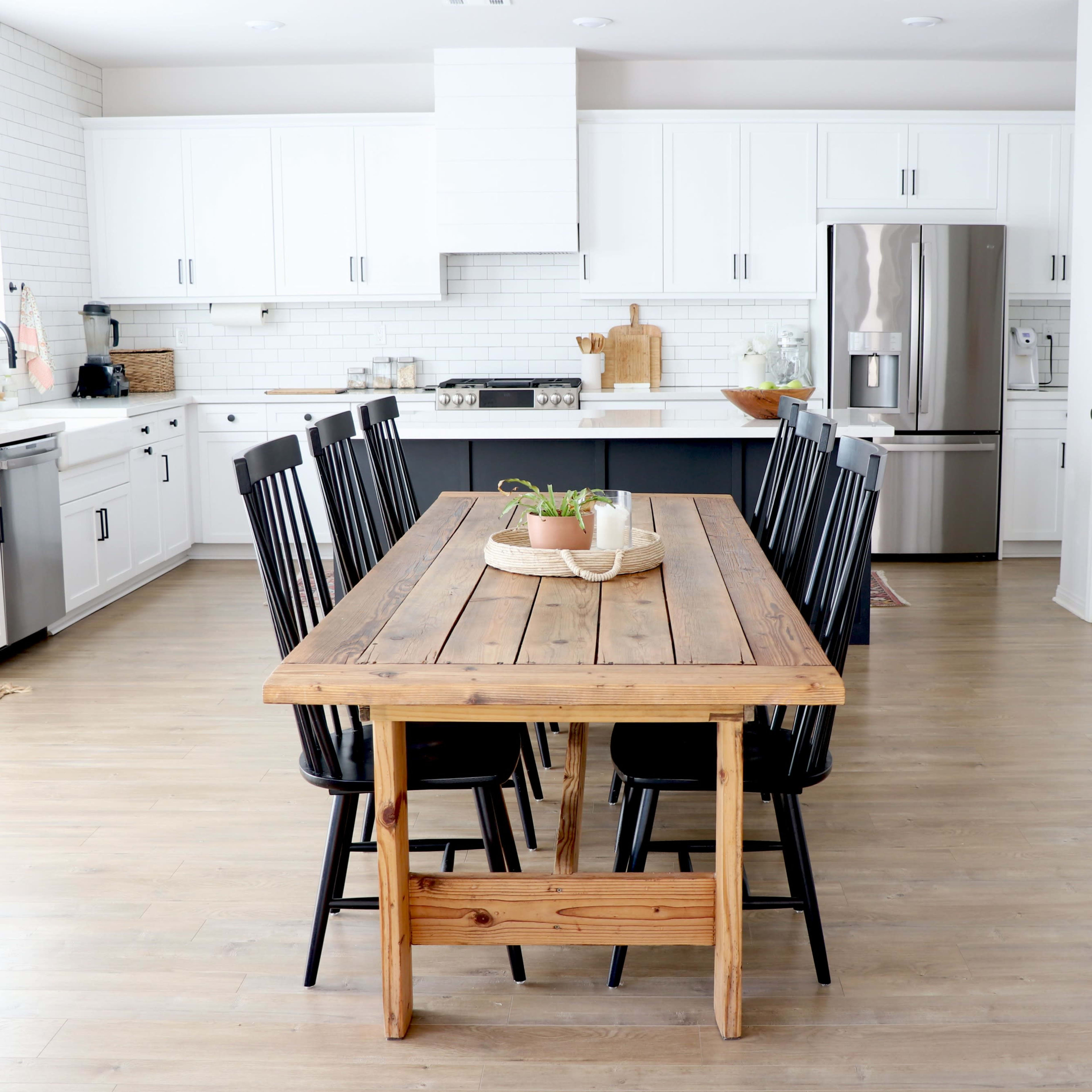 A large wooden dining table with black chairs is centered in a modern kitchen featuring white cabinets and stainless steel appliances.