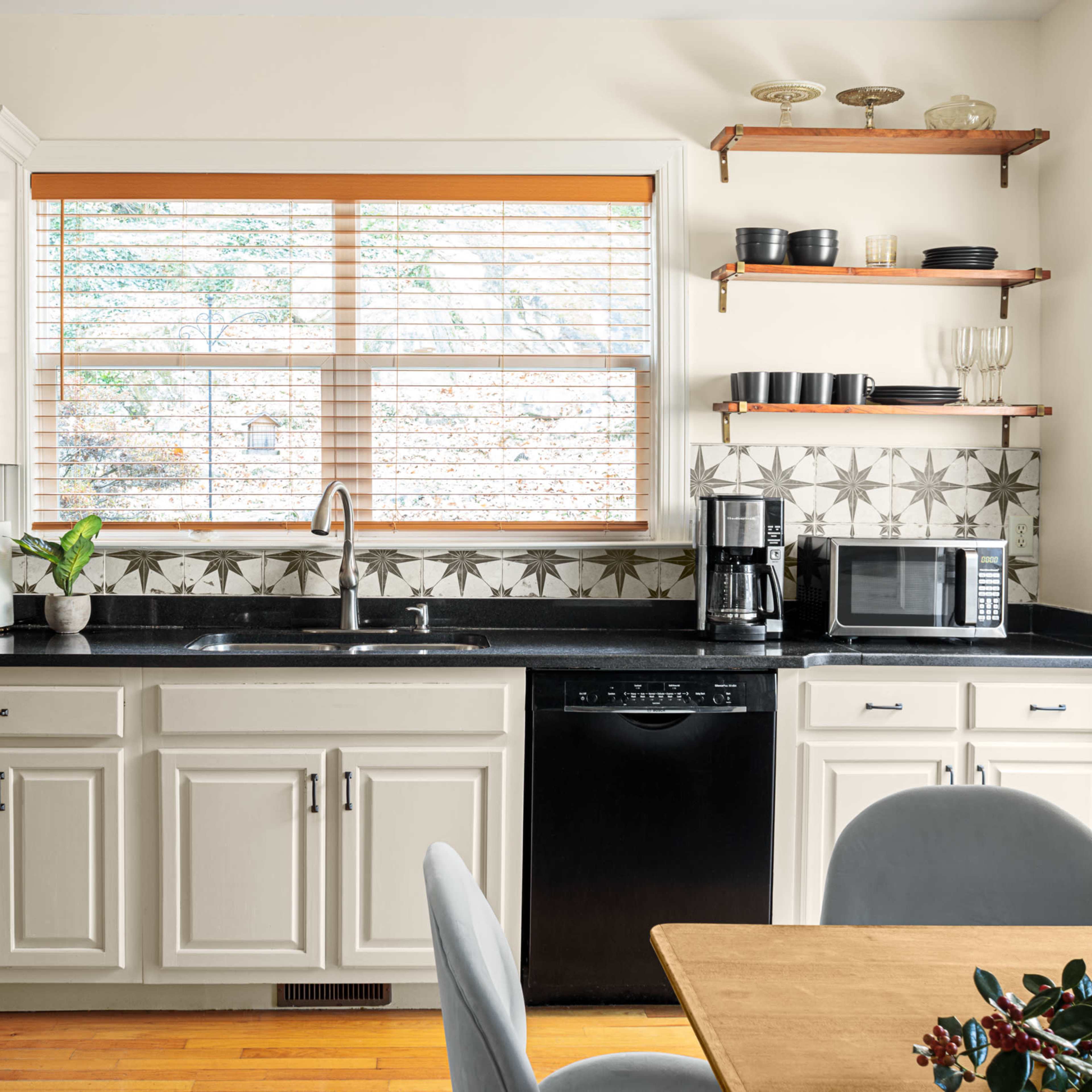A kitchen with white cabinets, a black countertop, a sink under a window, a microwave, a coffee maker, and a wooden dining table with gray chairs.