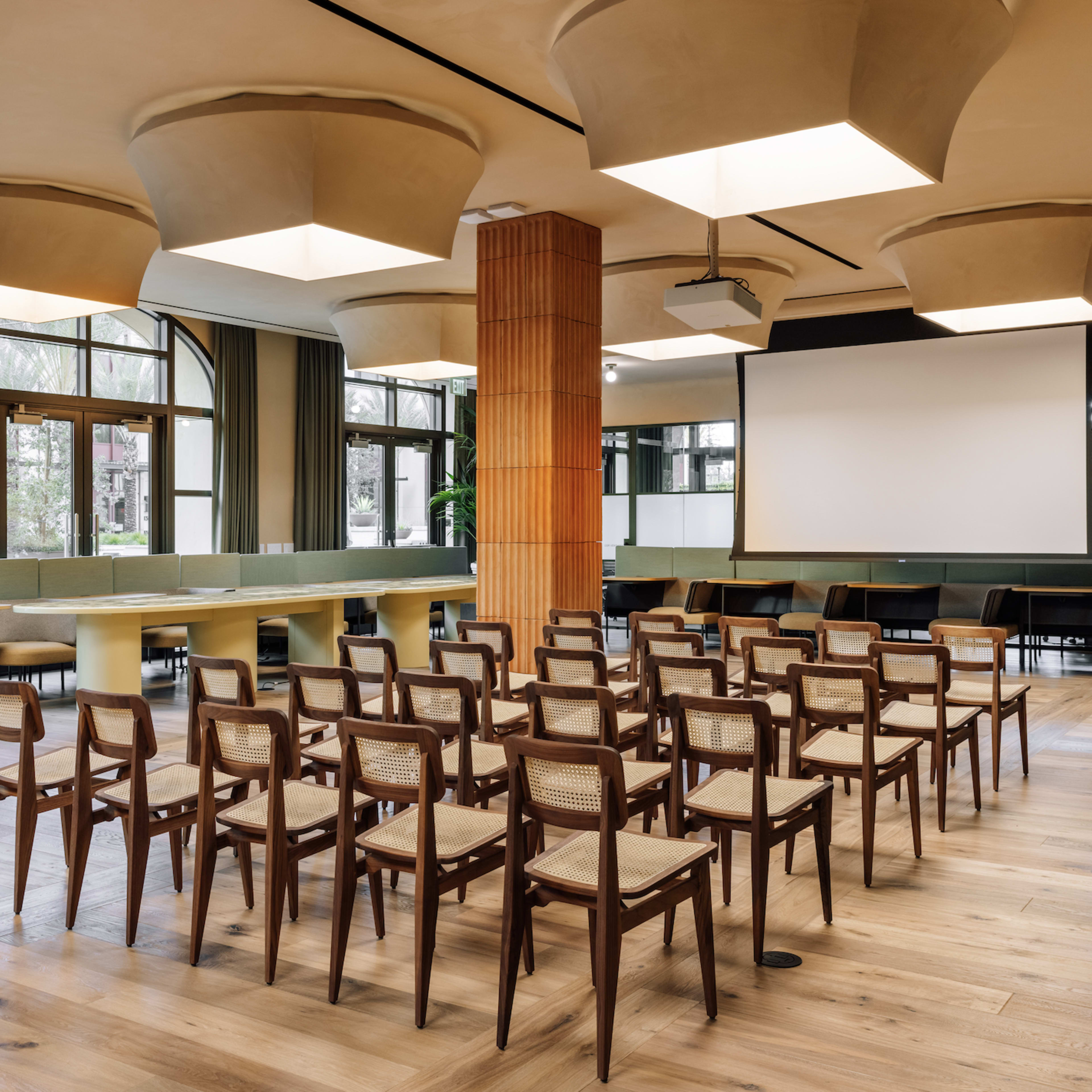 A spacious conference room with rows of wooden chairs, a large table, and a projector screen at one end.