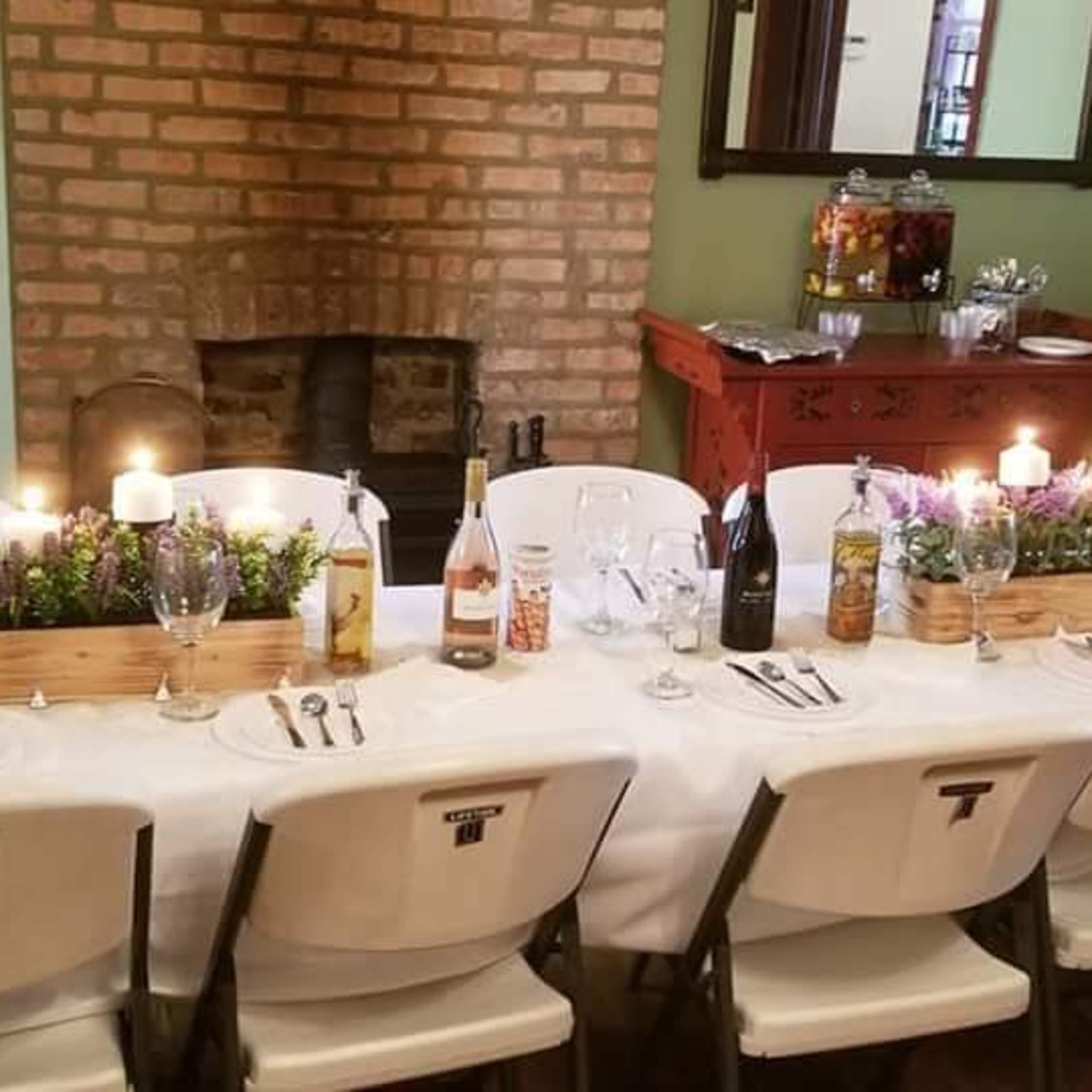 A long table is set for a meal with plates, glasses, and candles, arranged neatly in a room featuring a brick wall and a decorative sideboard.
