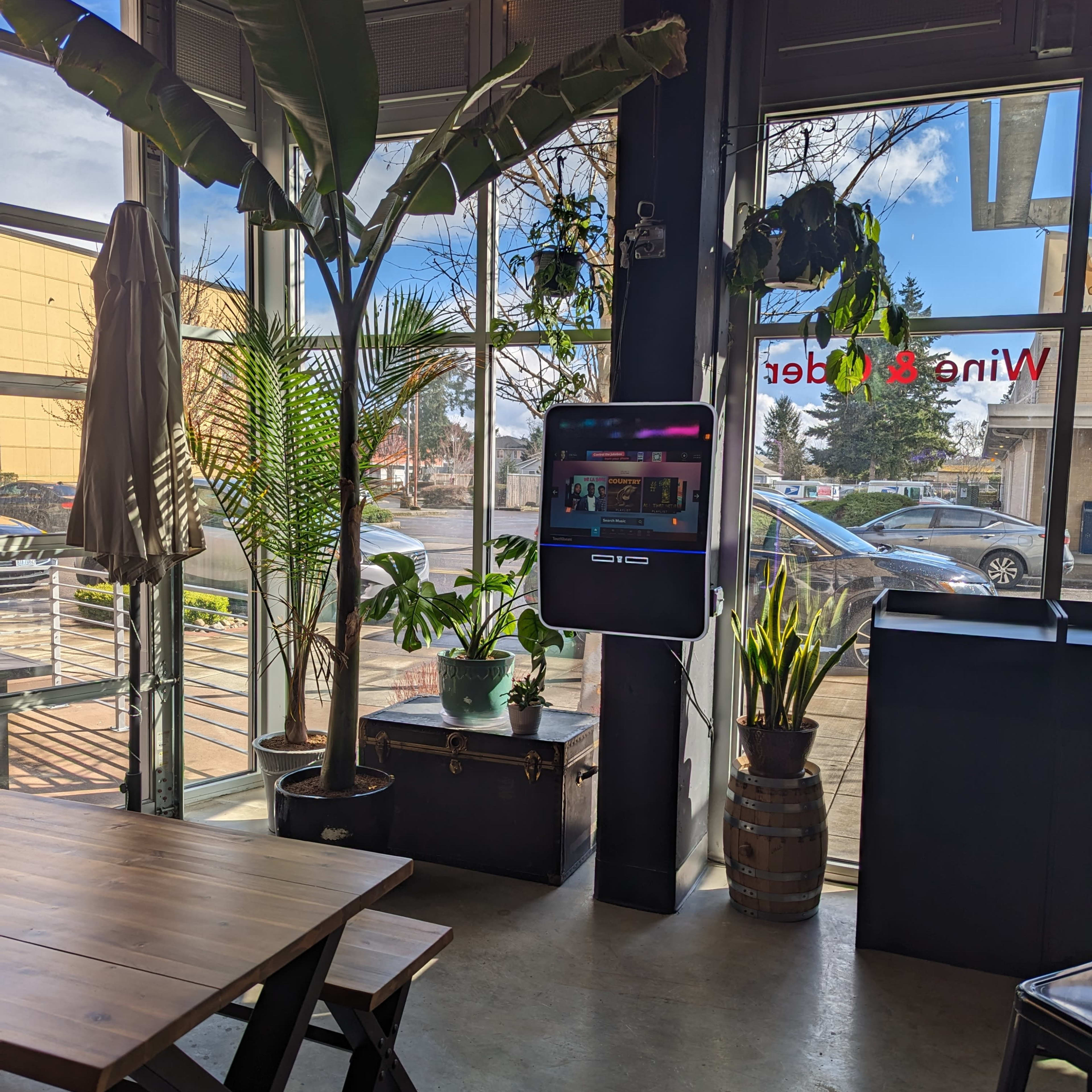 A bright interior of a cafe with large windows, potted plants, a table, and a digital kiosk on the wall.