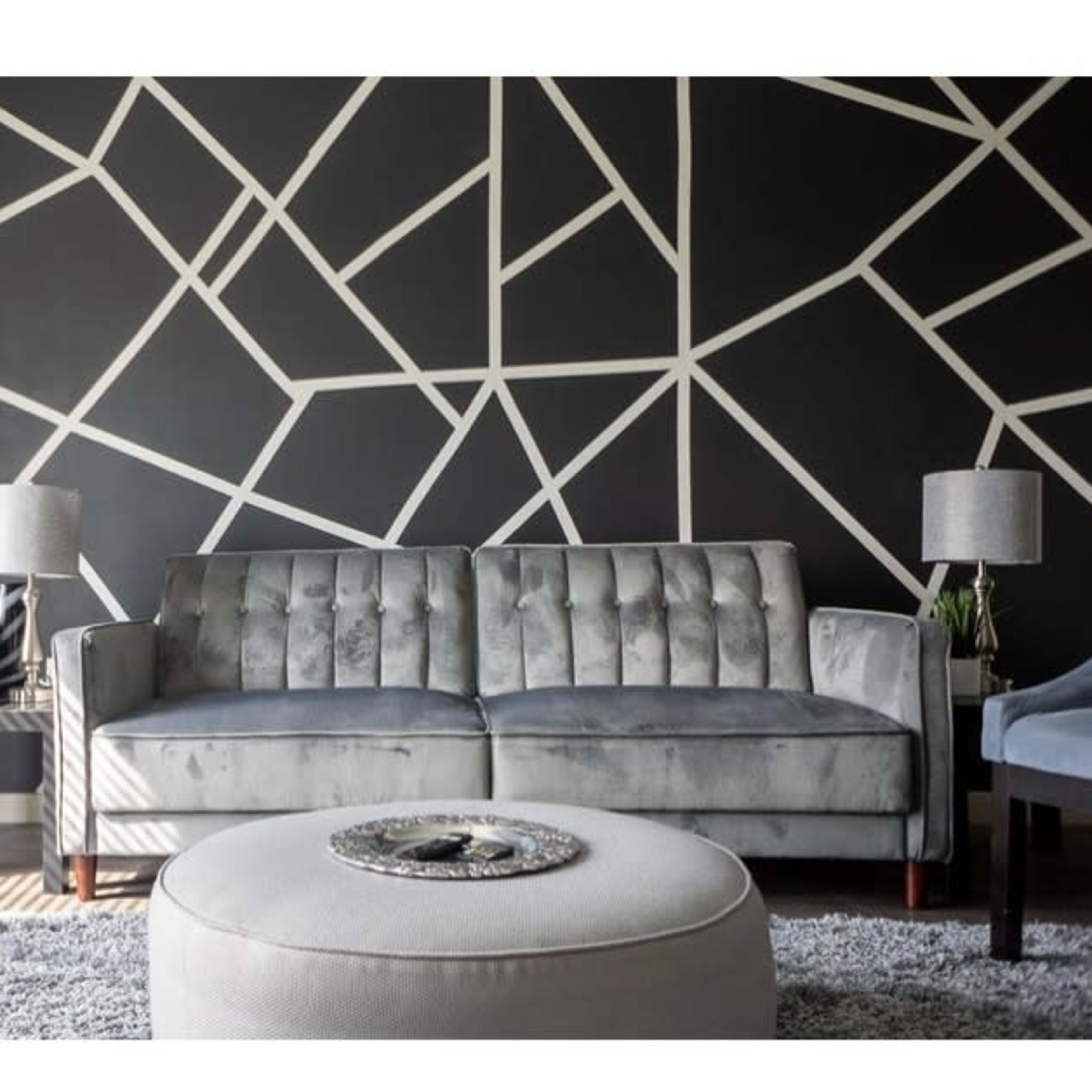 A modern living room with a gray velvet sofa, a round ottoman, and a geometric black and white accent wall.