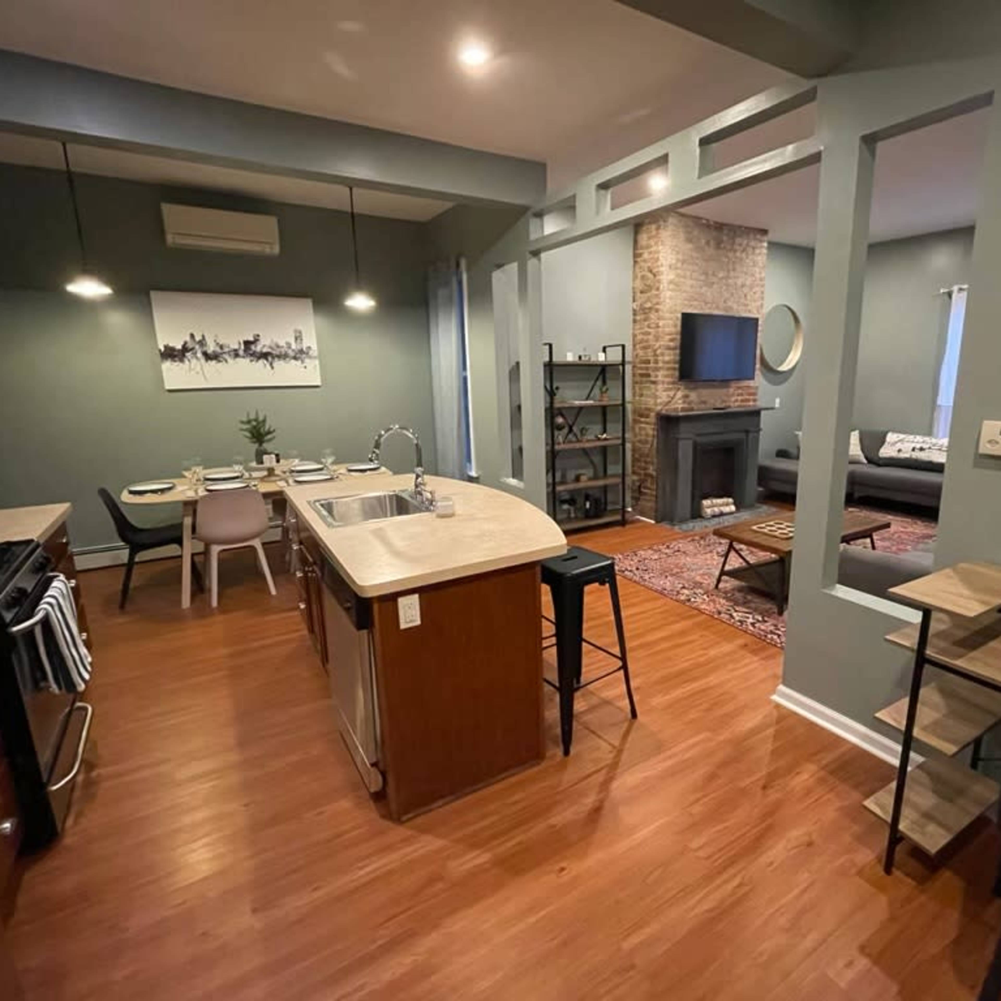 A modern open-concept kitchen and living area feature wood flooring, a stone fireplace, and a dining set.