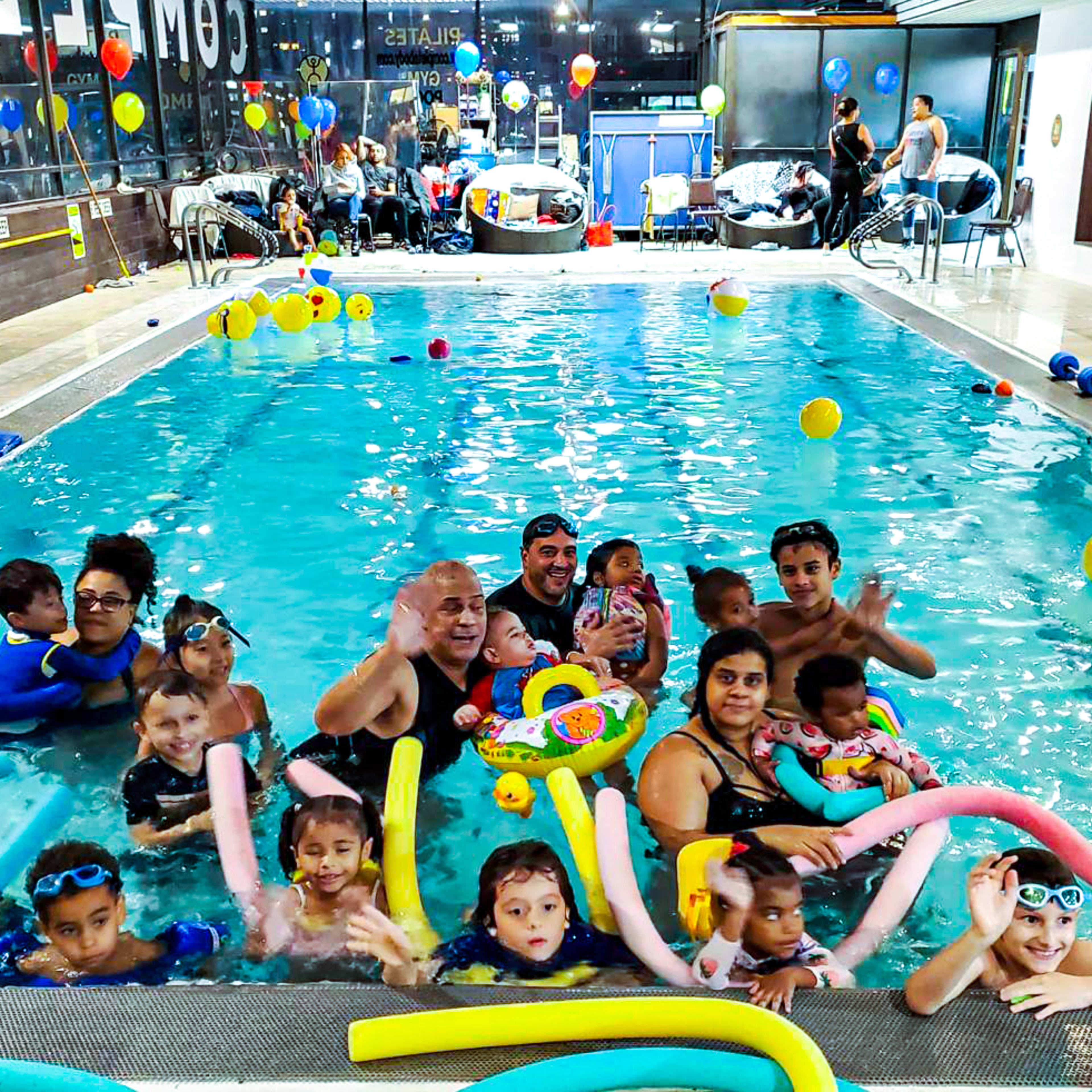 A large group of children and adults are playing in a pool filled with colorful inflatable toys and swim noodles.