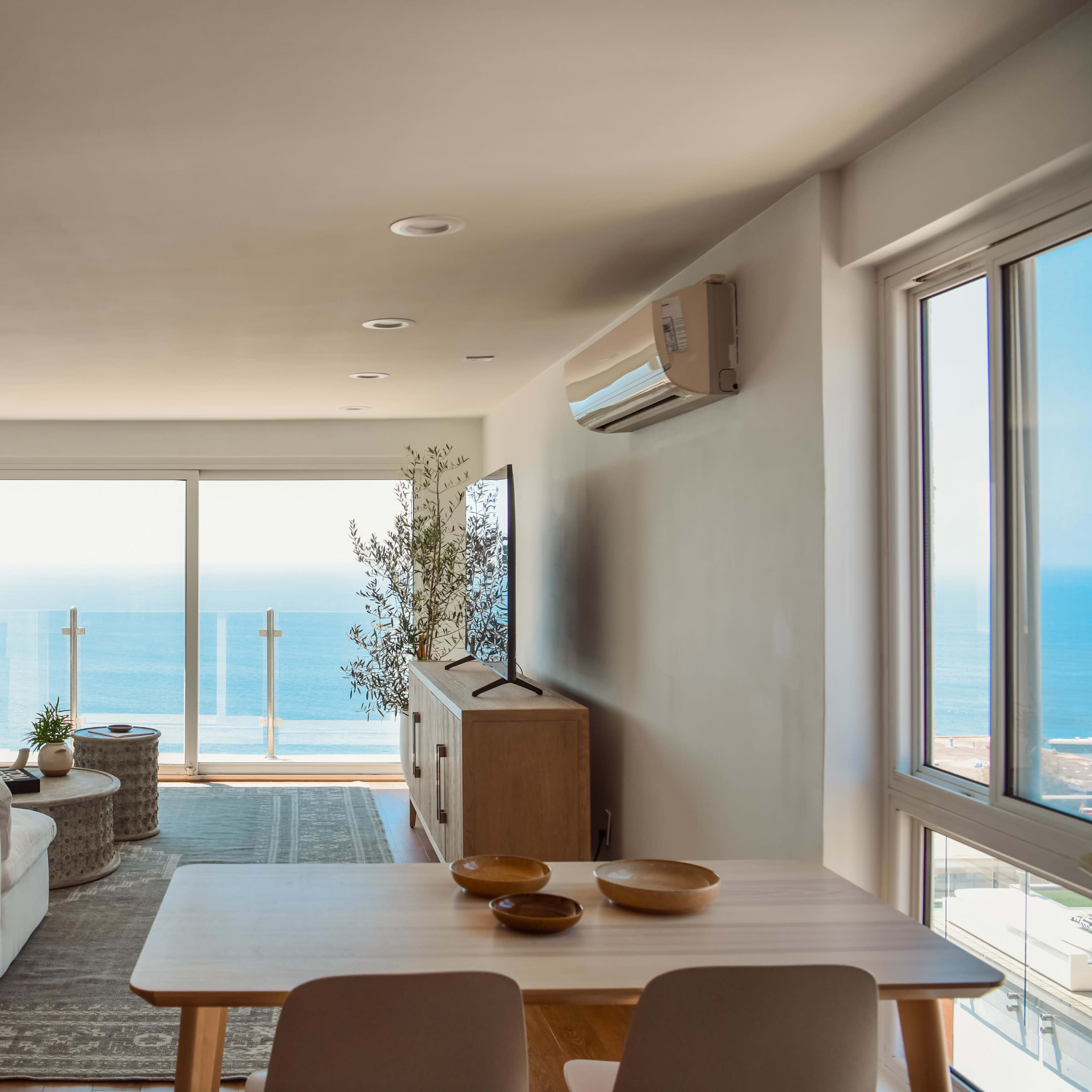 A bright living room with a large window overlooking the ocean, featuring a sofa, a coffee table, and a dining table set for four.