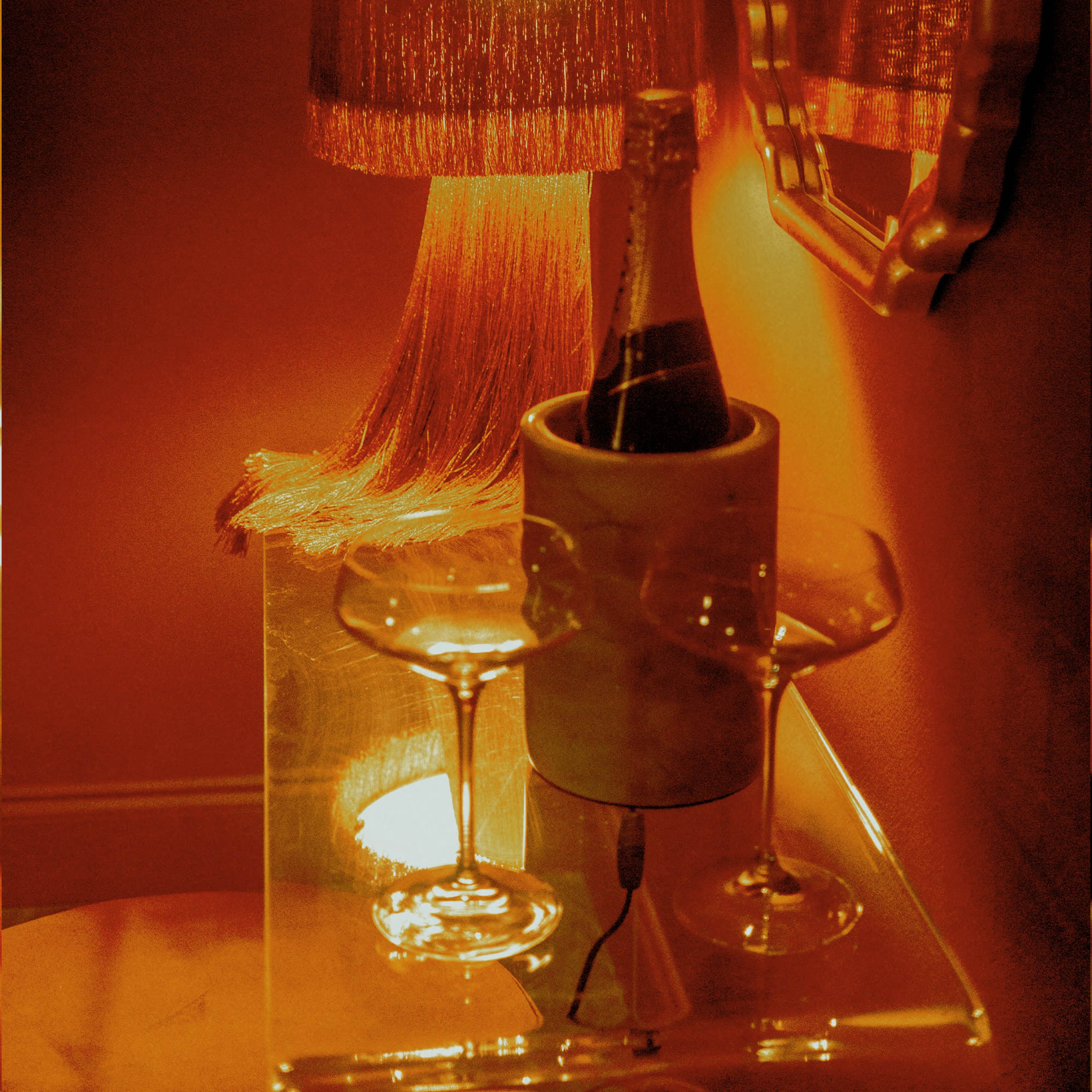 A lamp with a fringed shade illuminates a small table holding a champagne bottle in an ice bucket alongside two empty glasses.