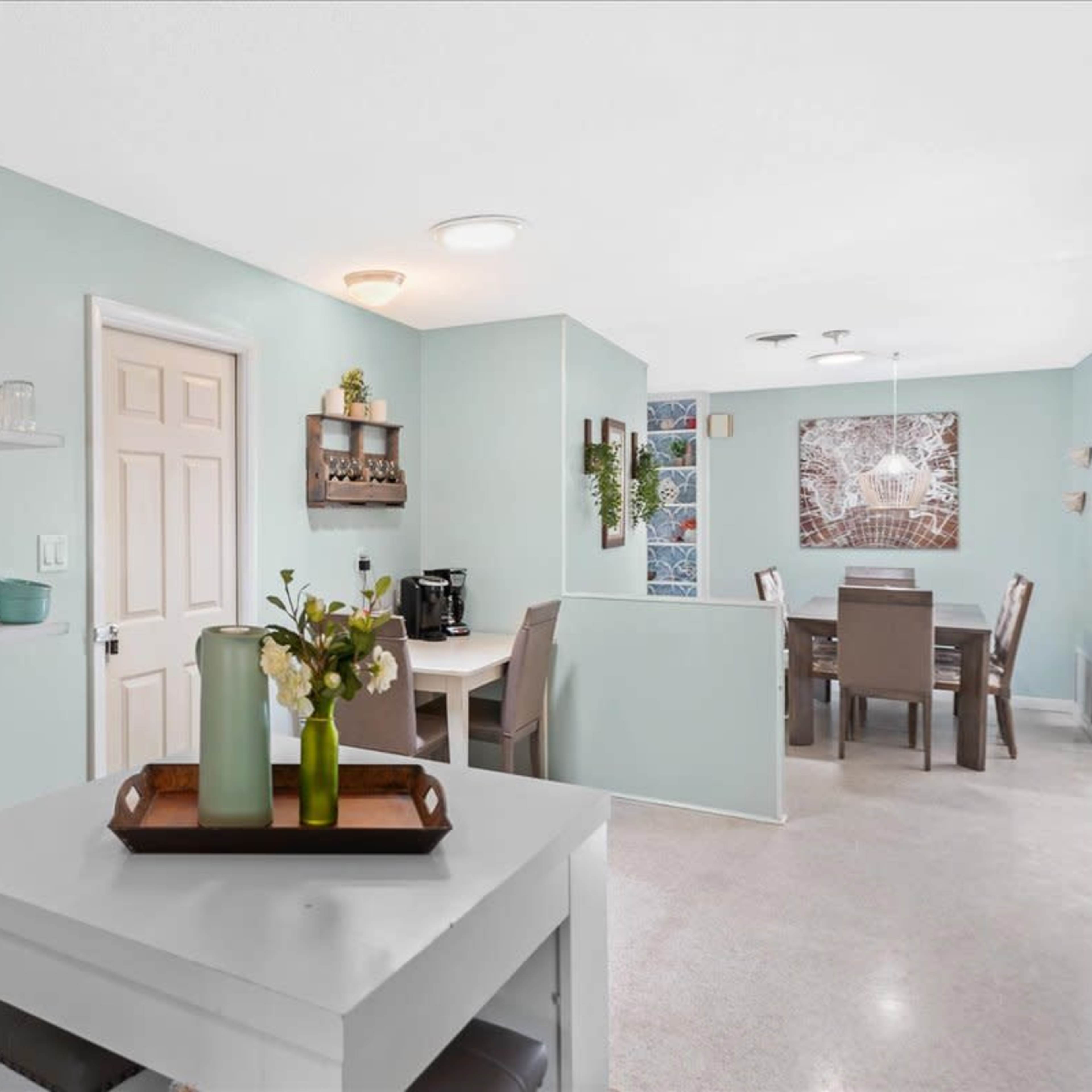 A light blue kitchen area with shelves holding glassware and dishes, a small table, and a dining area in the background.