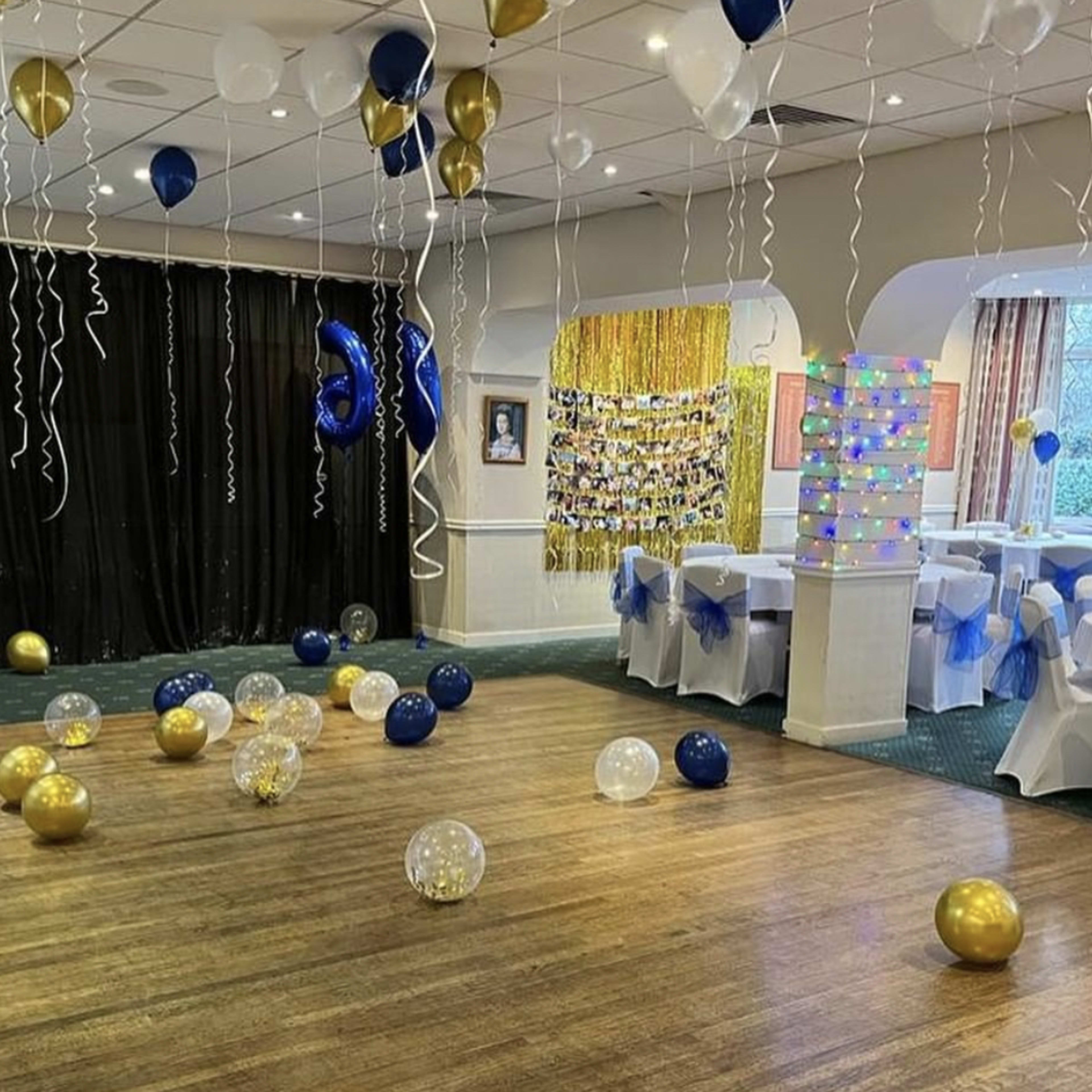 The room is decorated for a celebration, featuring scattered blue, gold, and clear balloons on the floor, with tables dressed in white and blue at the back.