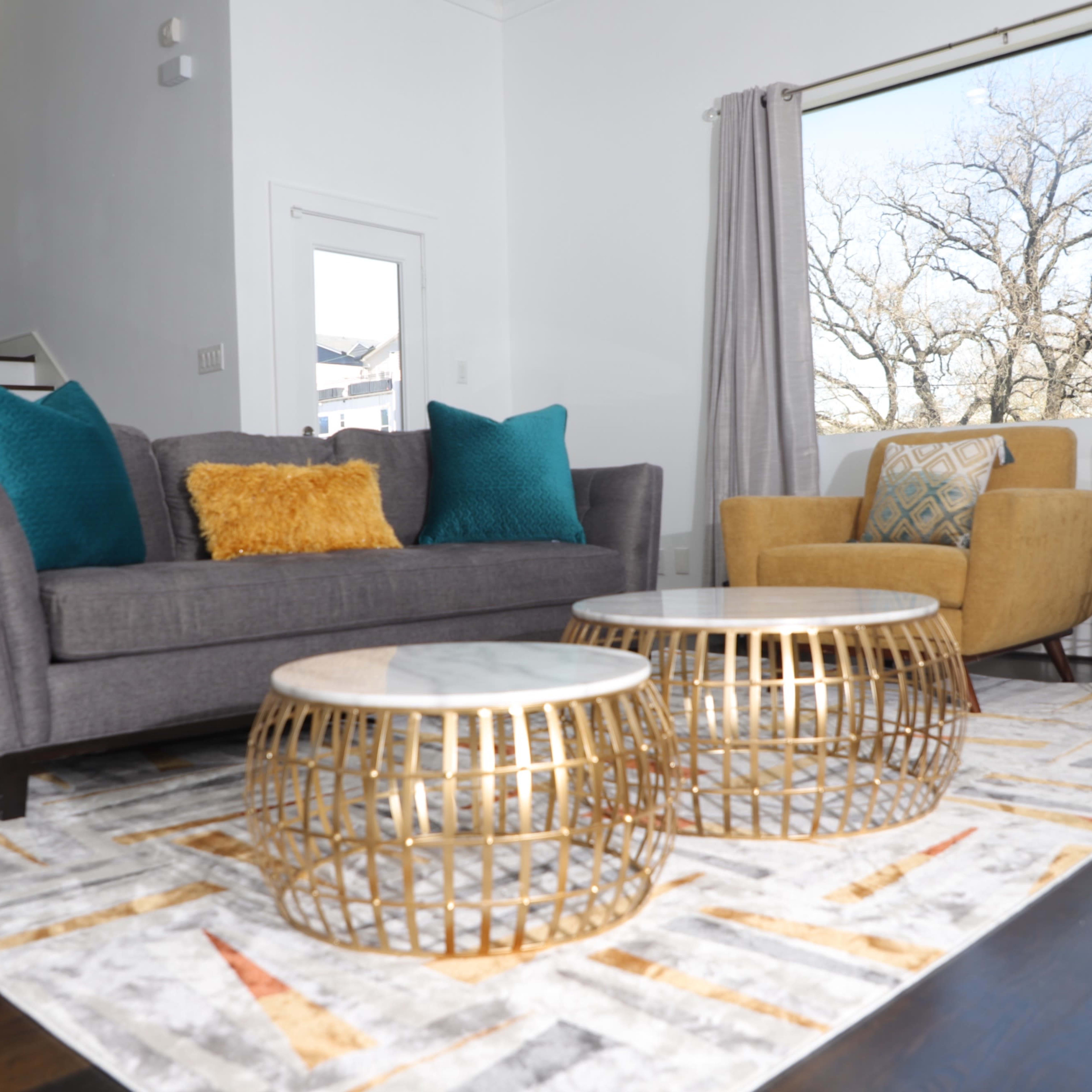 A modern living room with a gray sofa, an armchair, two decorative gold tables, and a patterned rug, all set against a large window.