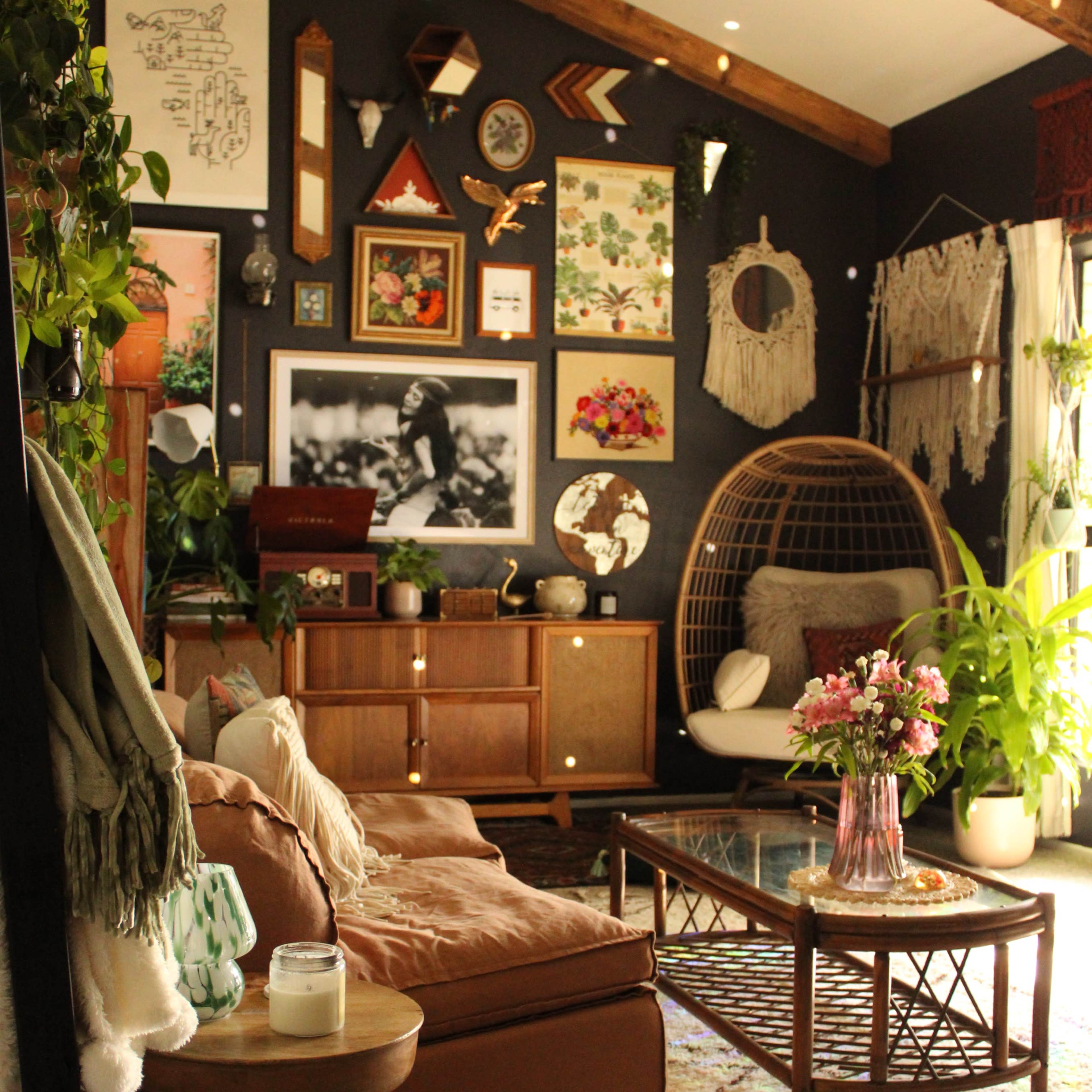 A cozy living room with a dark wall, a mix of decorative plants, art pieces, and comfortable furniture, including a rattan chair and a wooden coffee table.