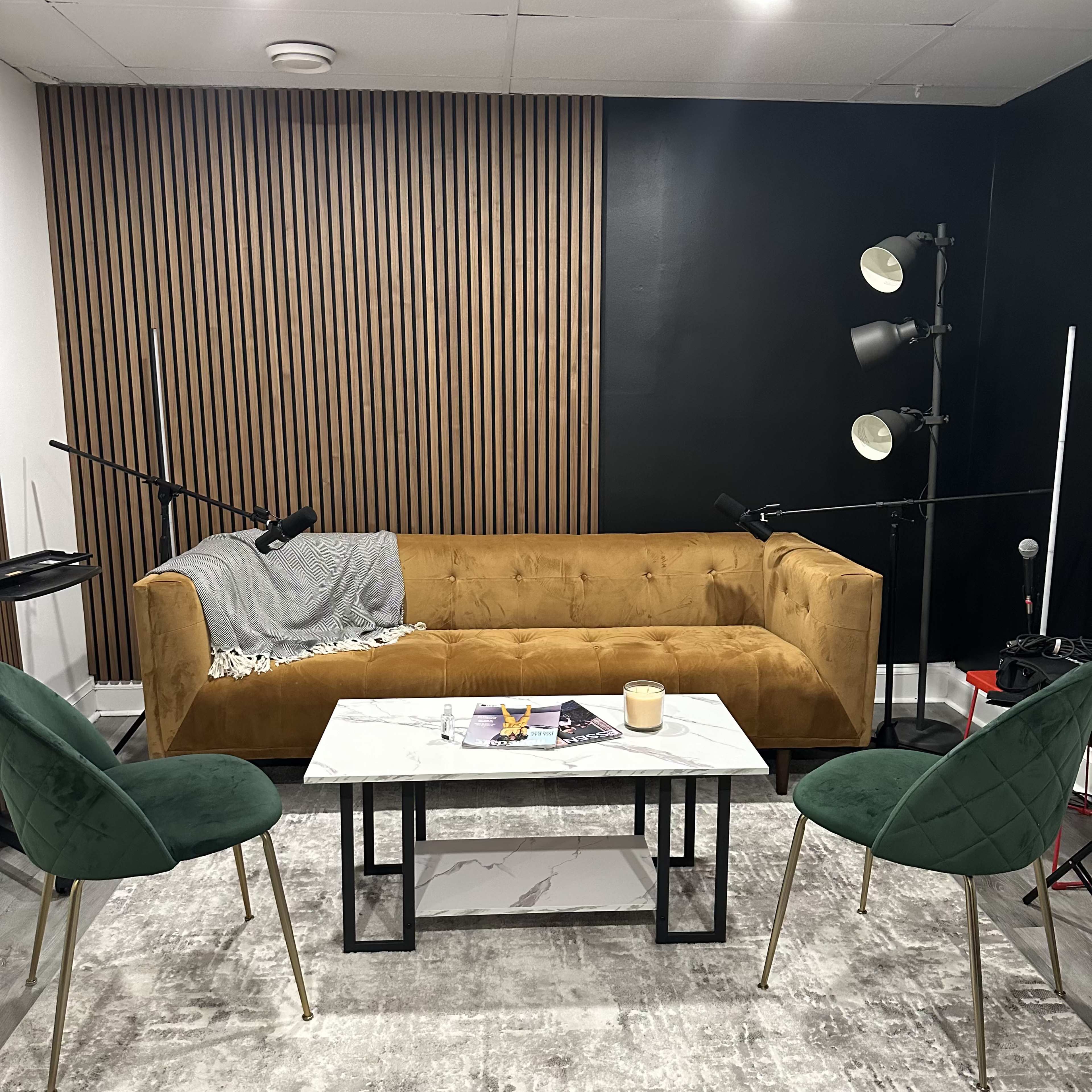 A living room setup with a brown velvet sofa, a marble coffee table, and two green chairs, all set against a backdrop of black and wooden paneling.