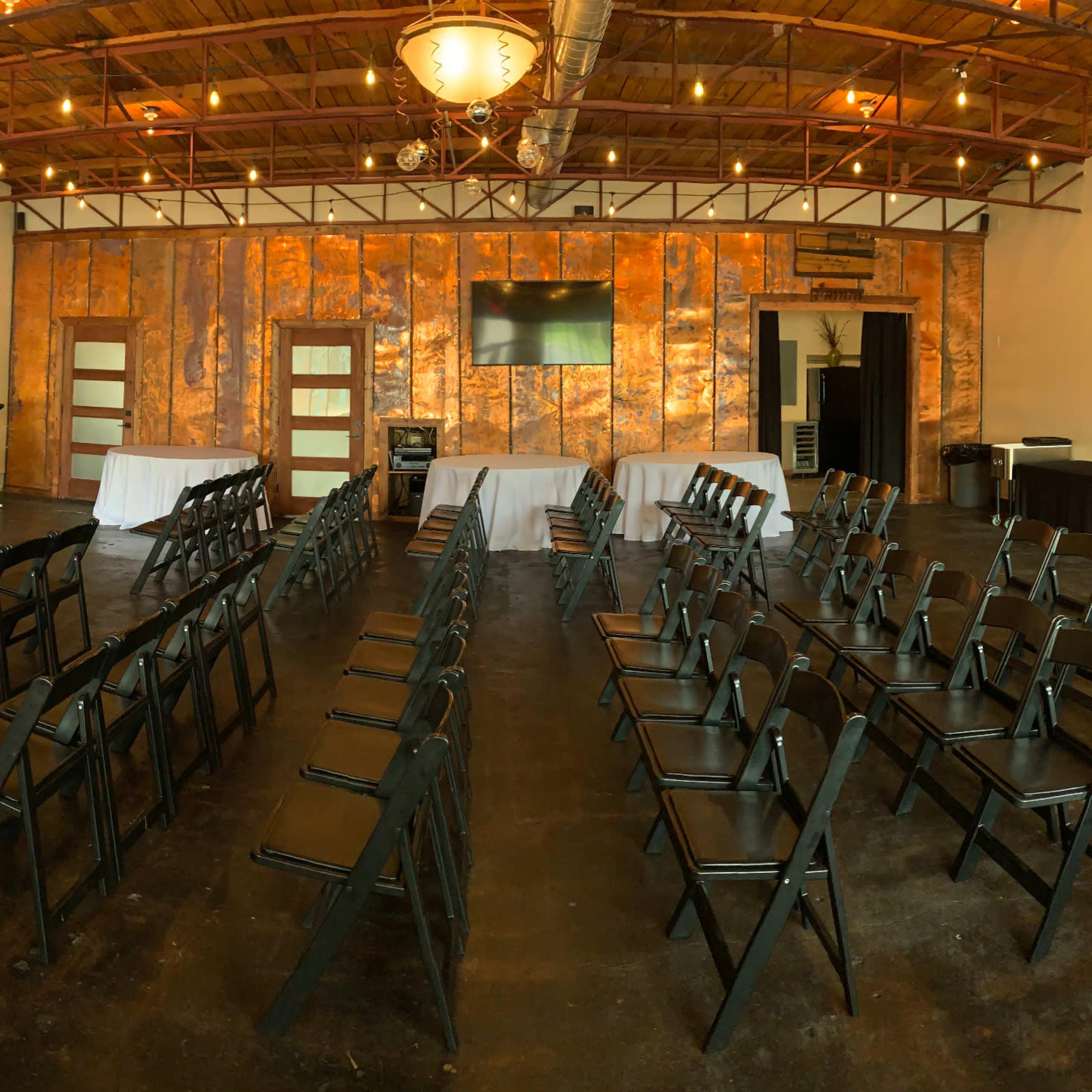 A spacious, well-lit event room set up with black chairs arranged in rows, round tables covered in white linens, and a buffet table along one wall.