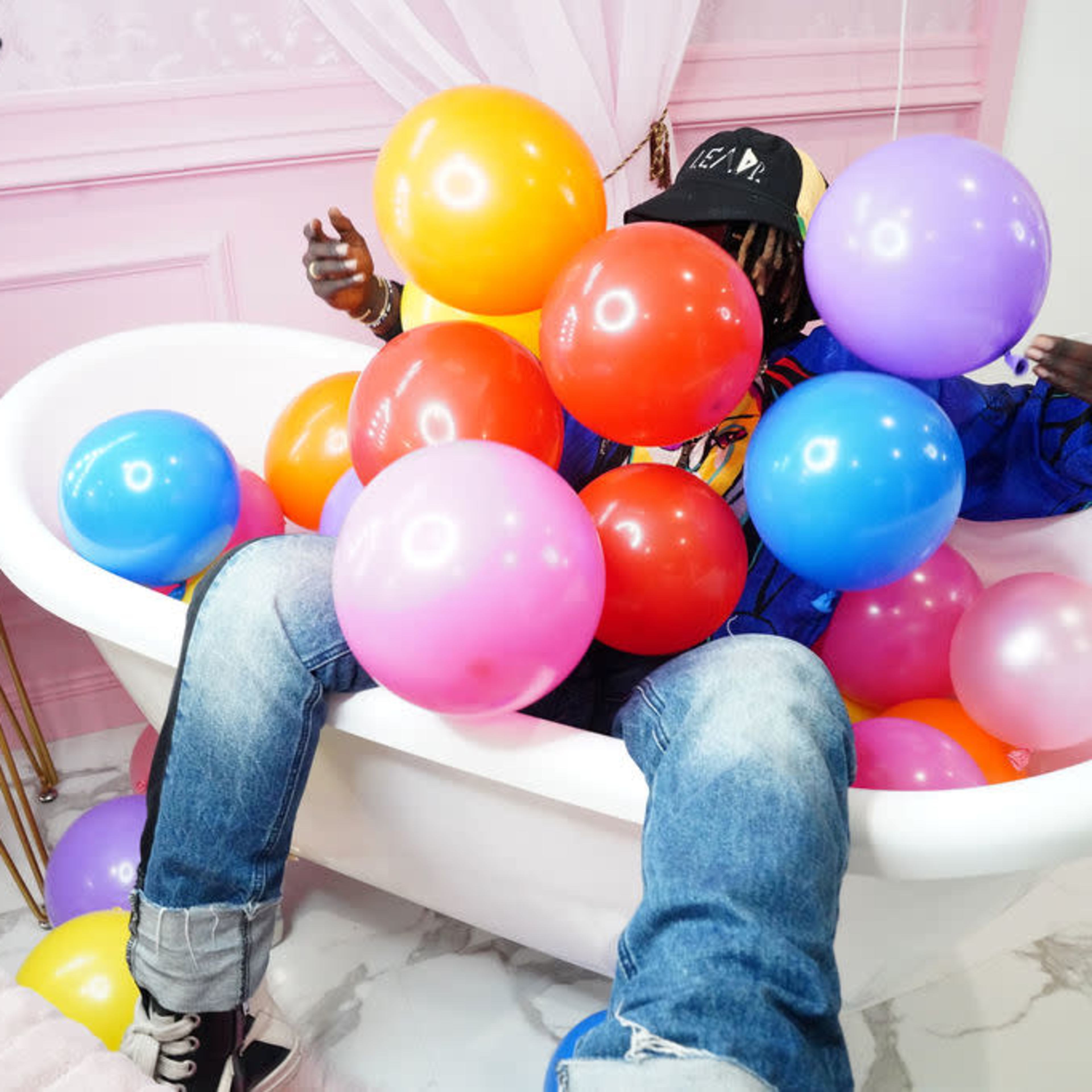 A person sitting in a bathtub filled with colorful balloons against a backdrop of pink walls and patterned decor.