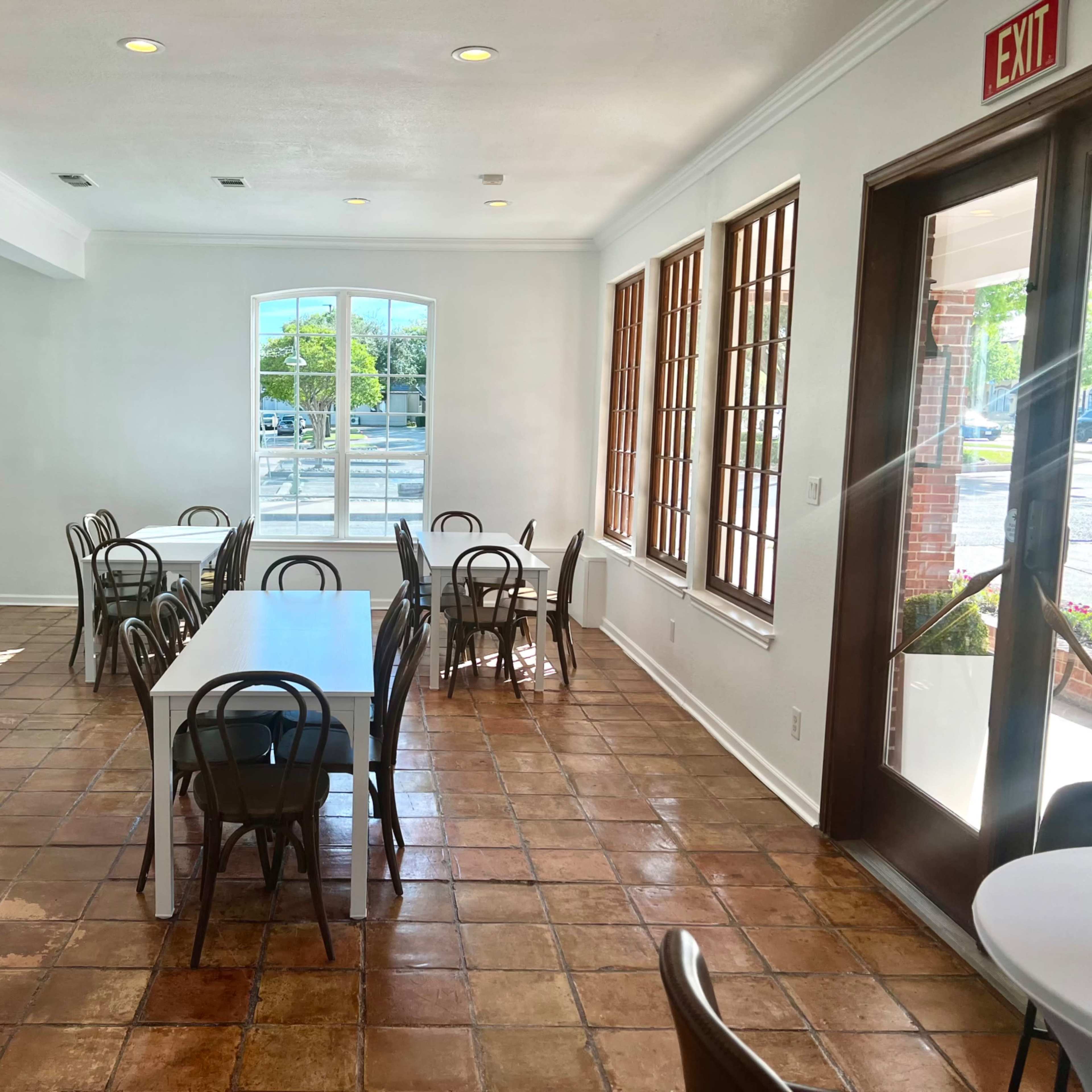A spacious dining area with multiple white tables and black chairs, featuring large windows that let in natural light.