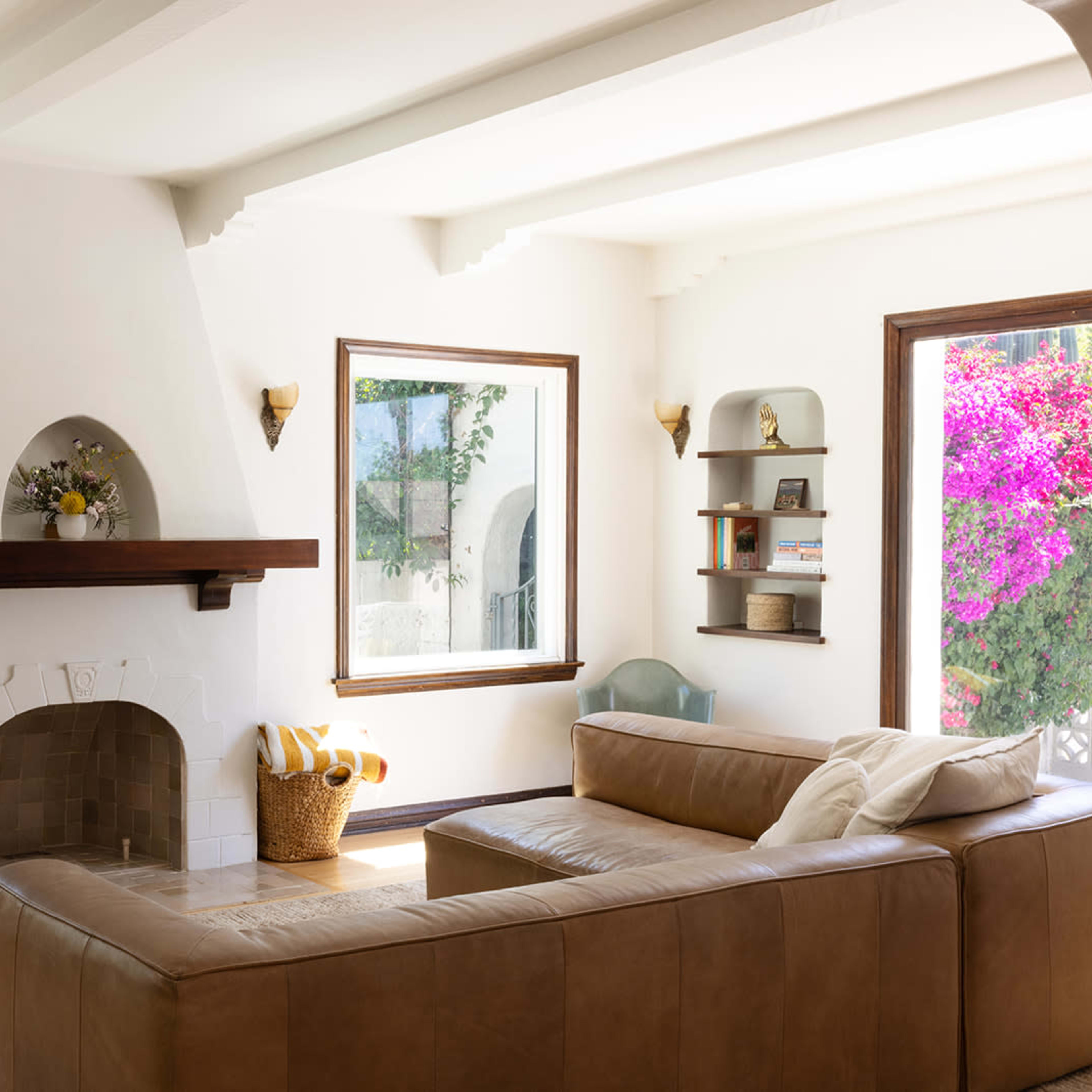 A spacious living room featuring a brown leather sectional sofa, an arched fireplace with a mantel, large windows, and shelves displaying decor, surrounded by greenery and pink flowers outside.