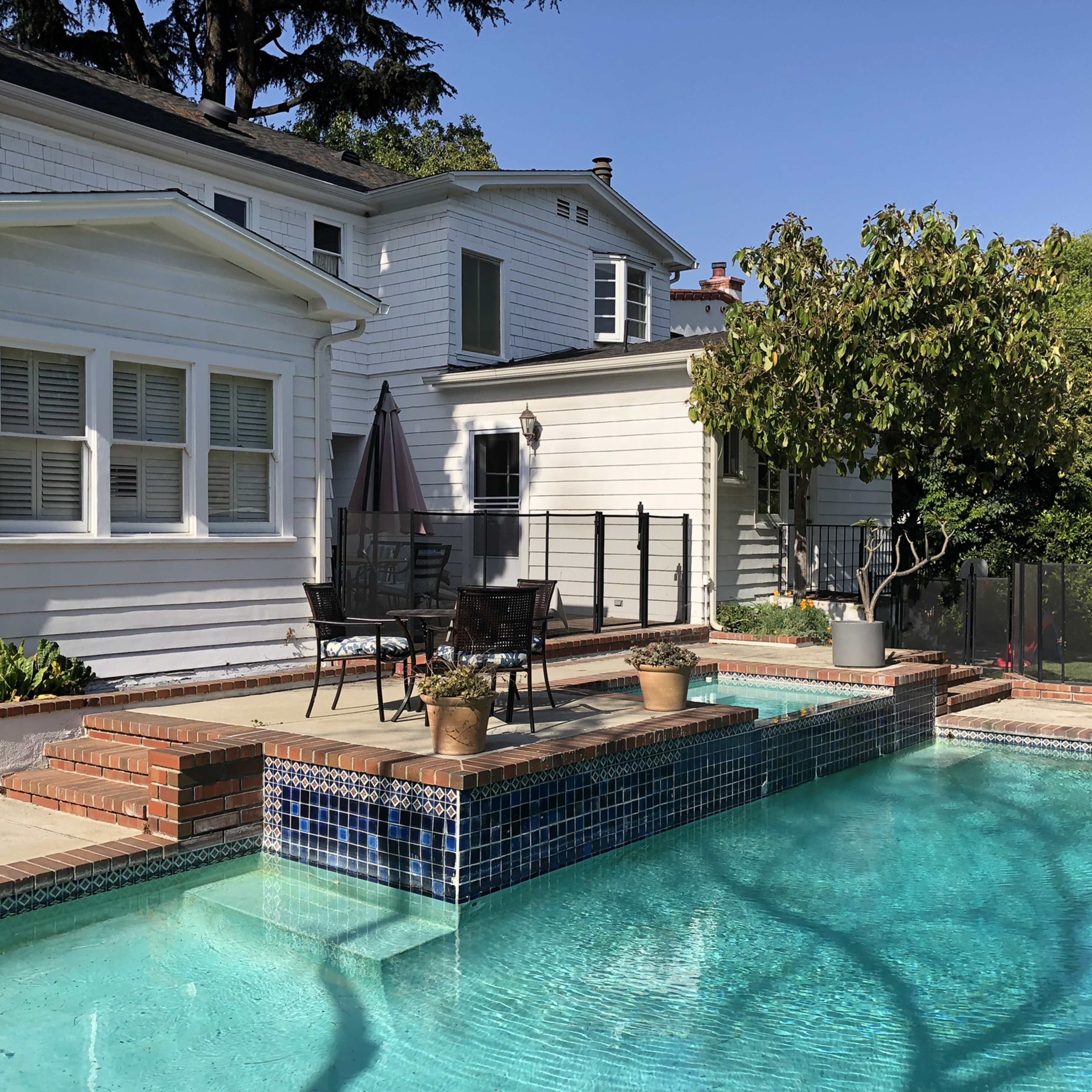 A clear swimming pool is lined with tiles and surrounded by a patio featuring potted plants and outdoor seating next to a white house with multiple sections.