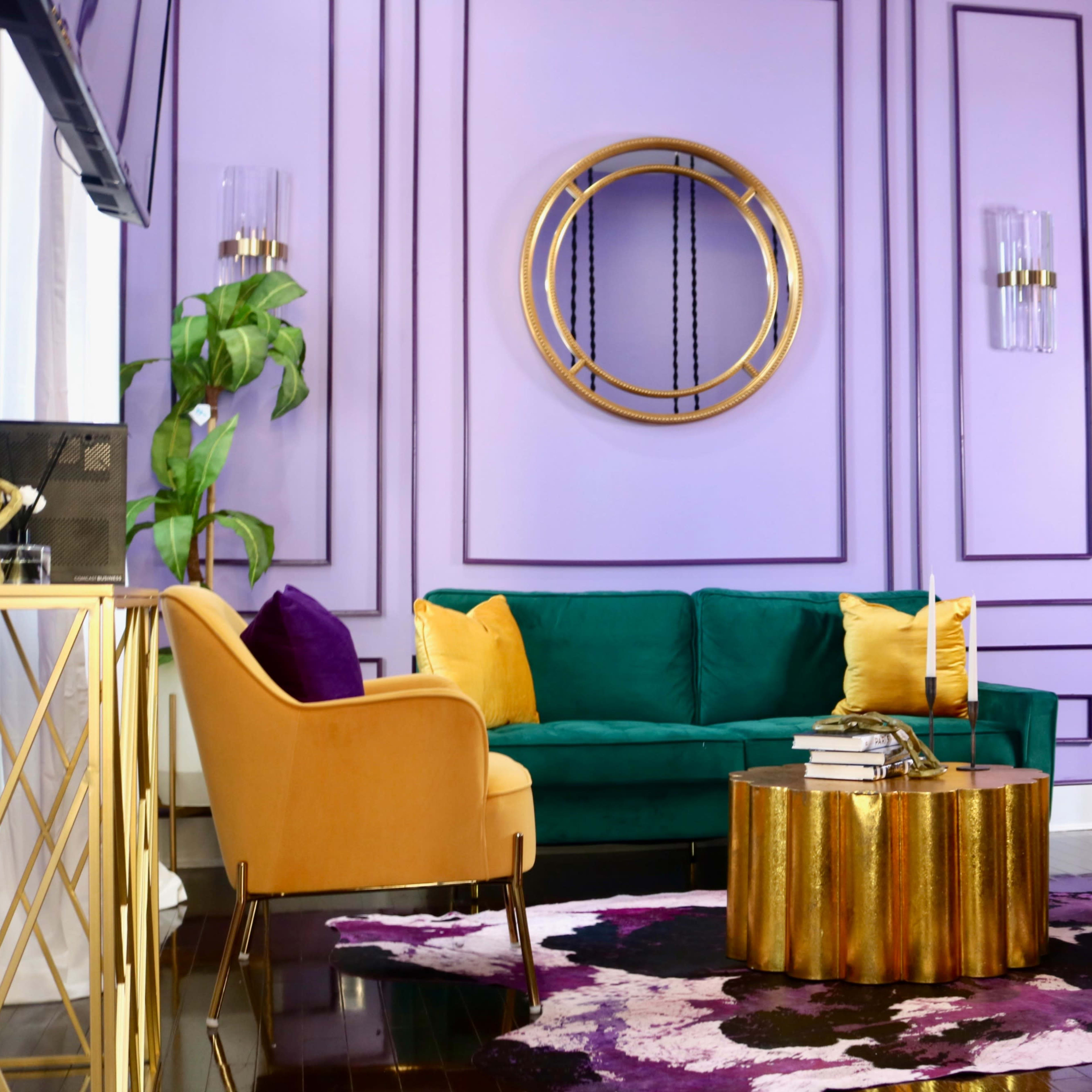 A vibrant living room features a green sofa and two light-colored chairs around a gold and wood coffee table, with purple and yellow accents against a lavender wall.