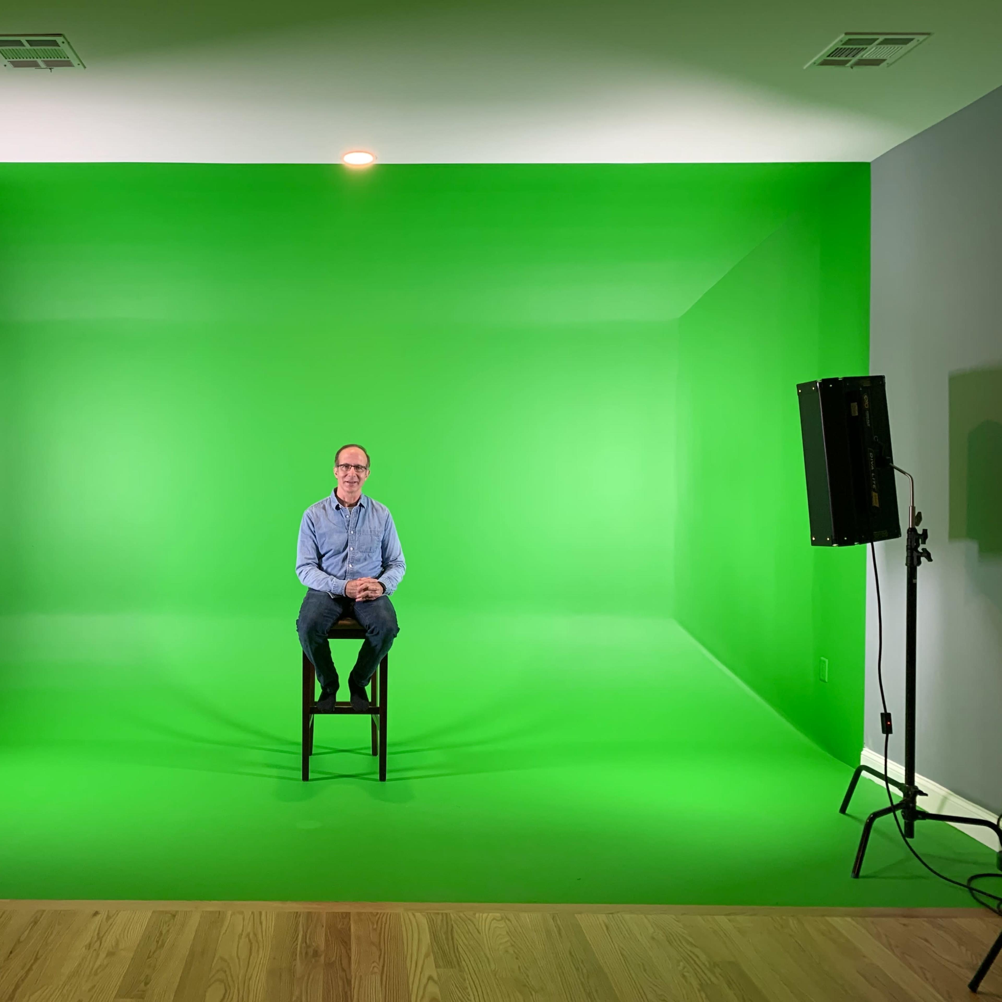 A man sits on a stool in front of a green screen, surrounded by two lights, in a spacious room with wooden flooring.