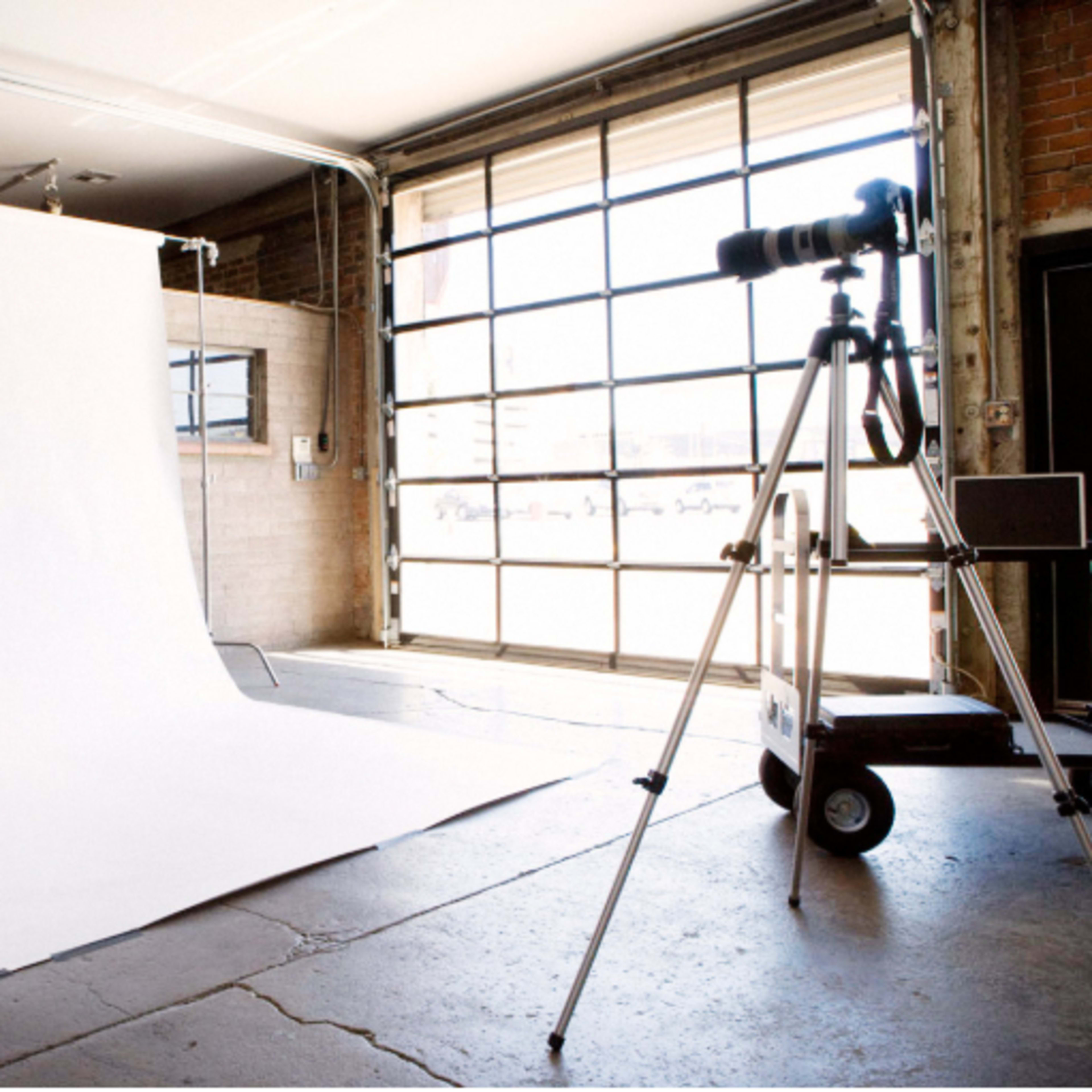 A camera is set up in a studio with a white backdrop and large garage-style windows.