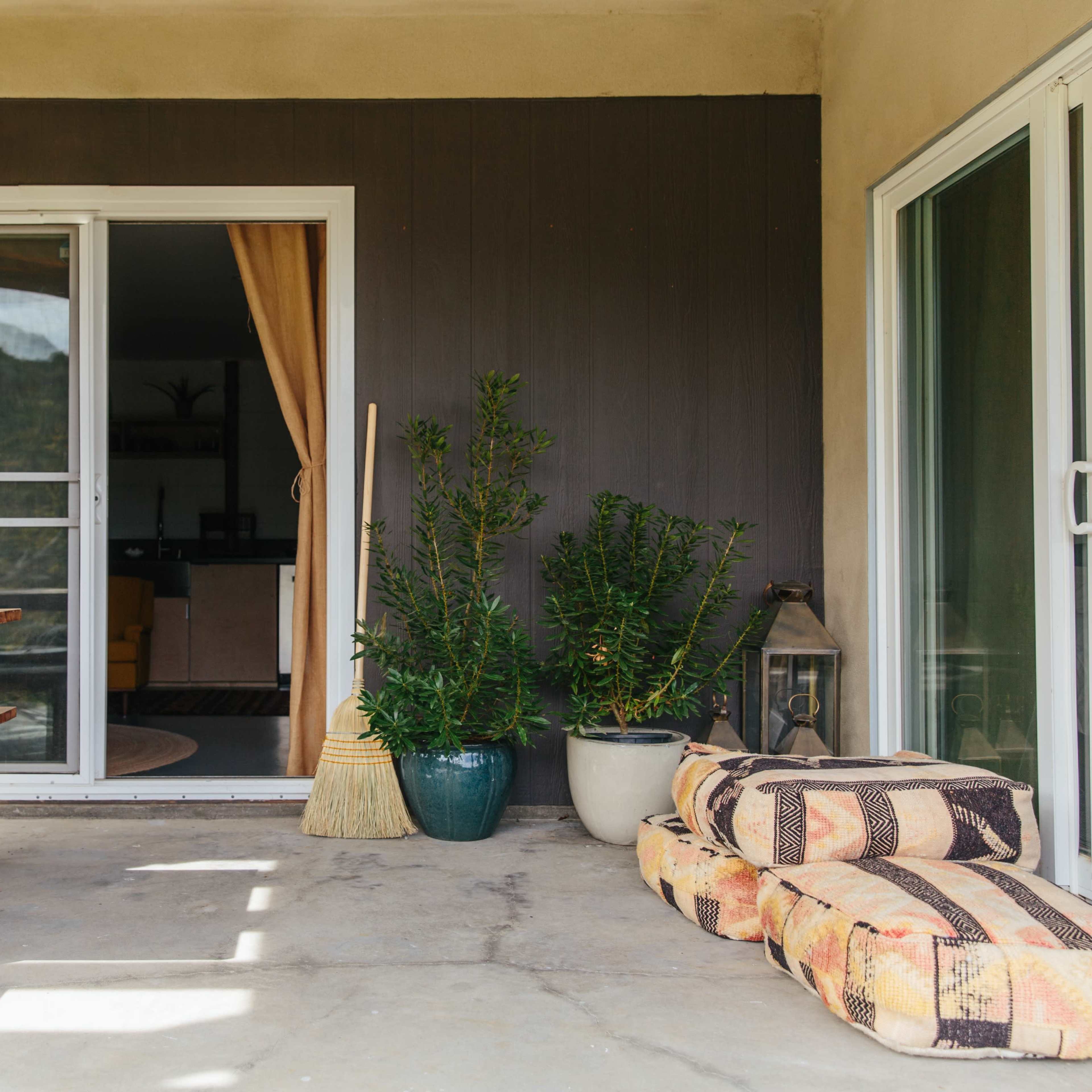 A patio area with a wooden table and benches, a broom, potted plants, and patterned cushions outside a sliding door.