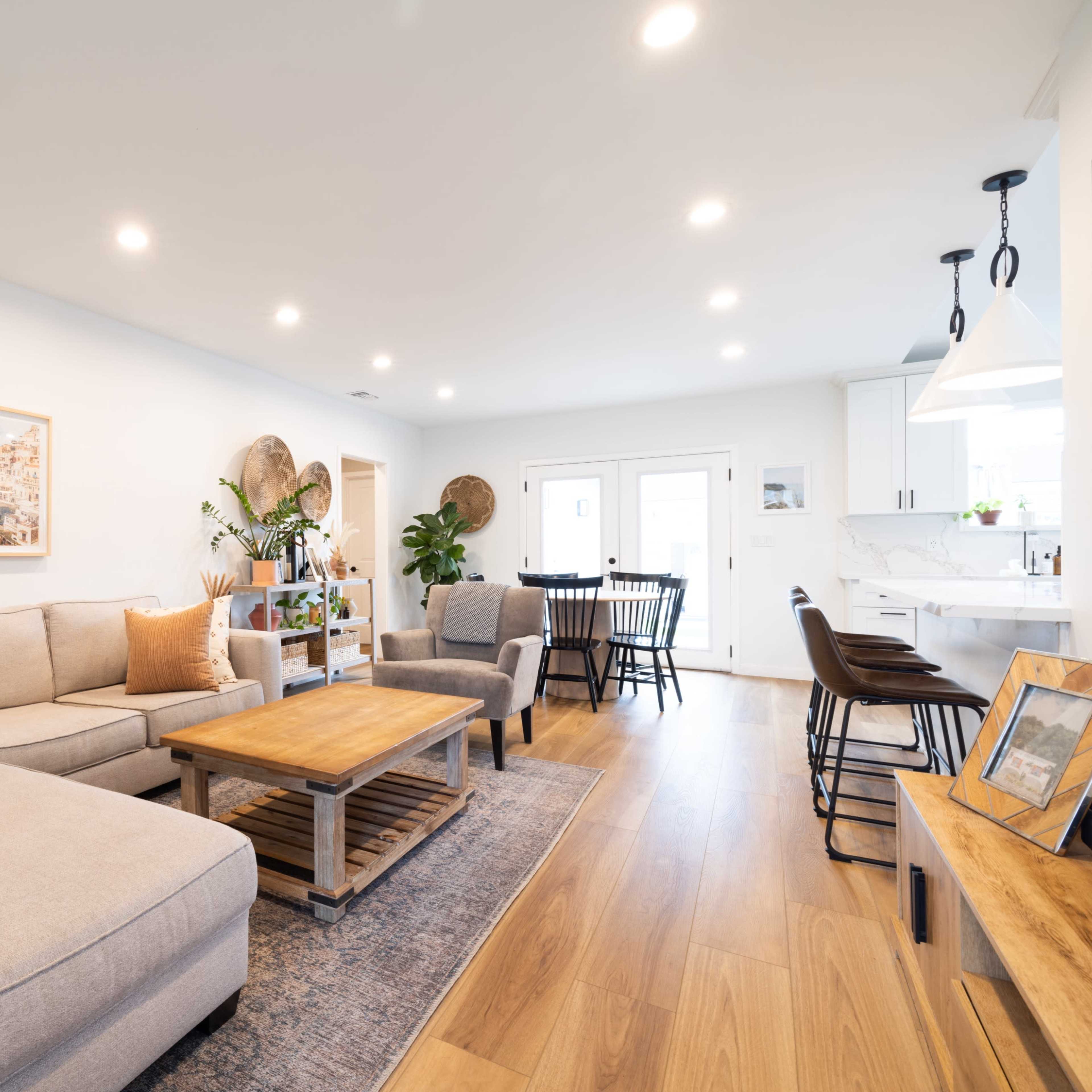 A light-filled living room features a sectional sofa, a wooden coffee table, and an adjacent dining area with a round table and chairs.