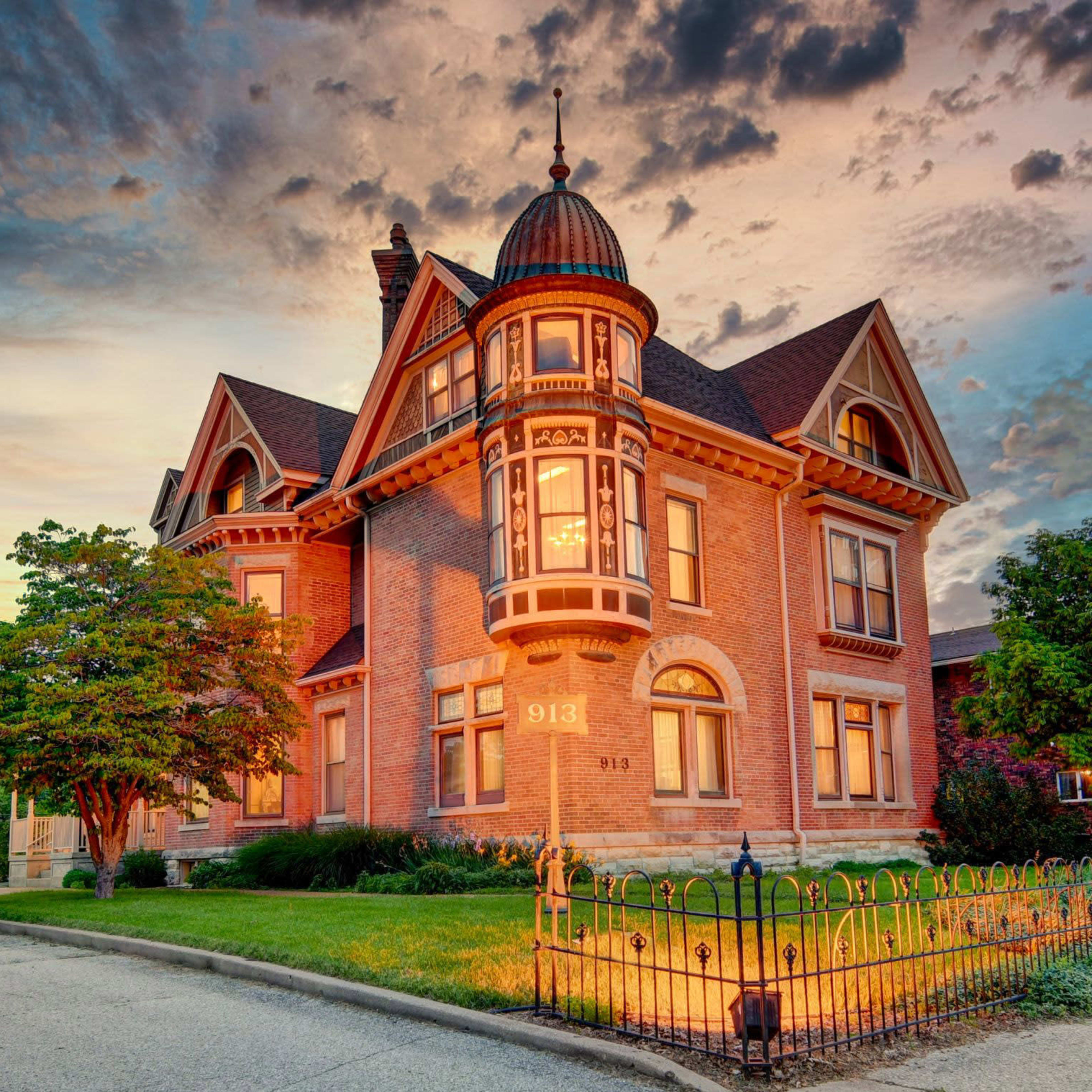 A historic brick mansion with a turret, surrounded by well-maintained greenery and a wrought-iron fence, under a colorful sky at dusk.