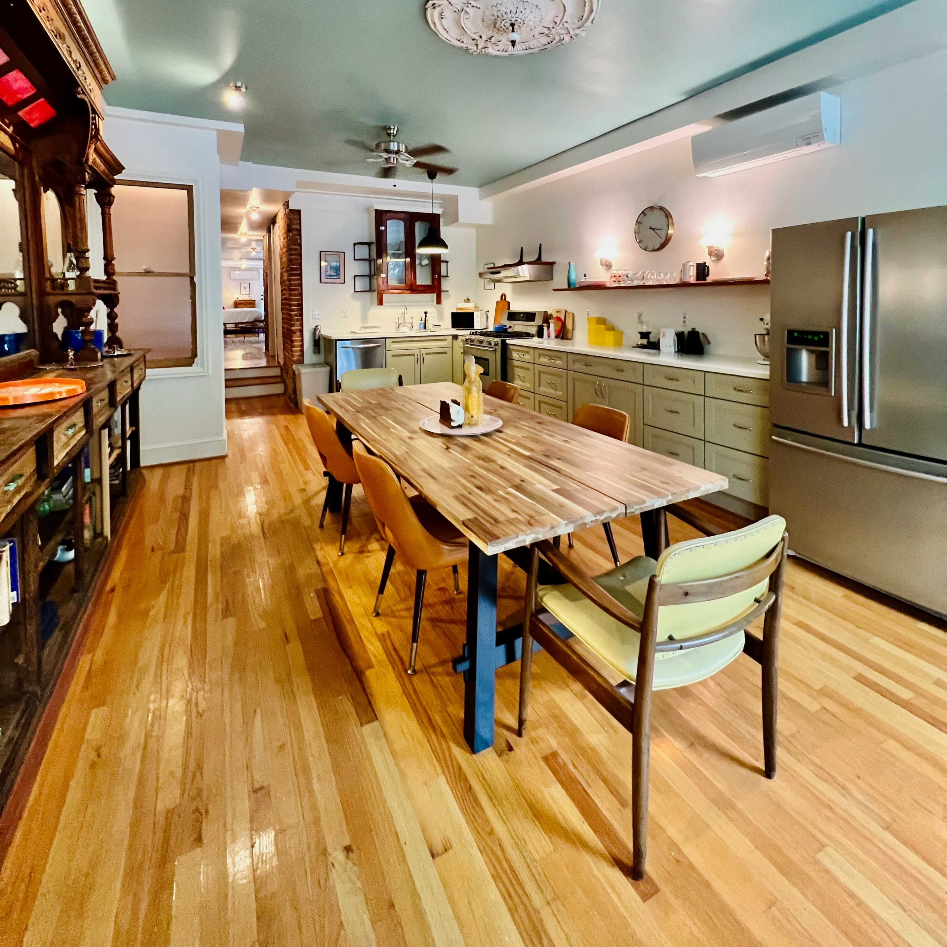 A well-lit kitchen with a wooden dining table surrounded by chairs, set against light-colored cabinetry and a stainless steel refrigerator.