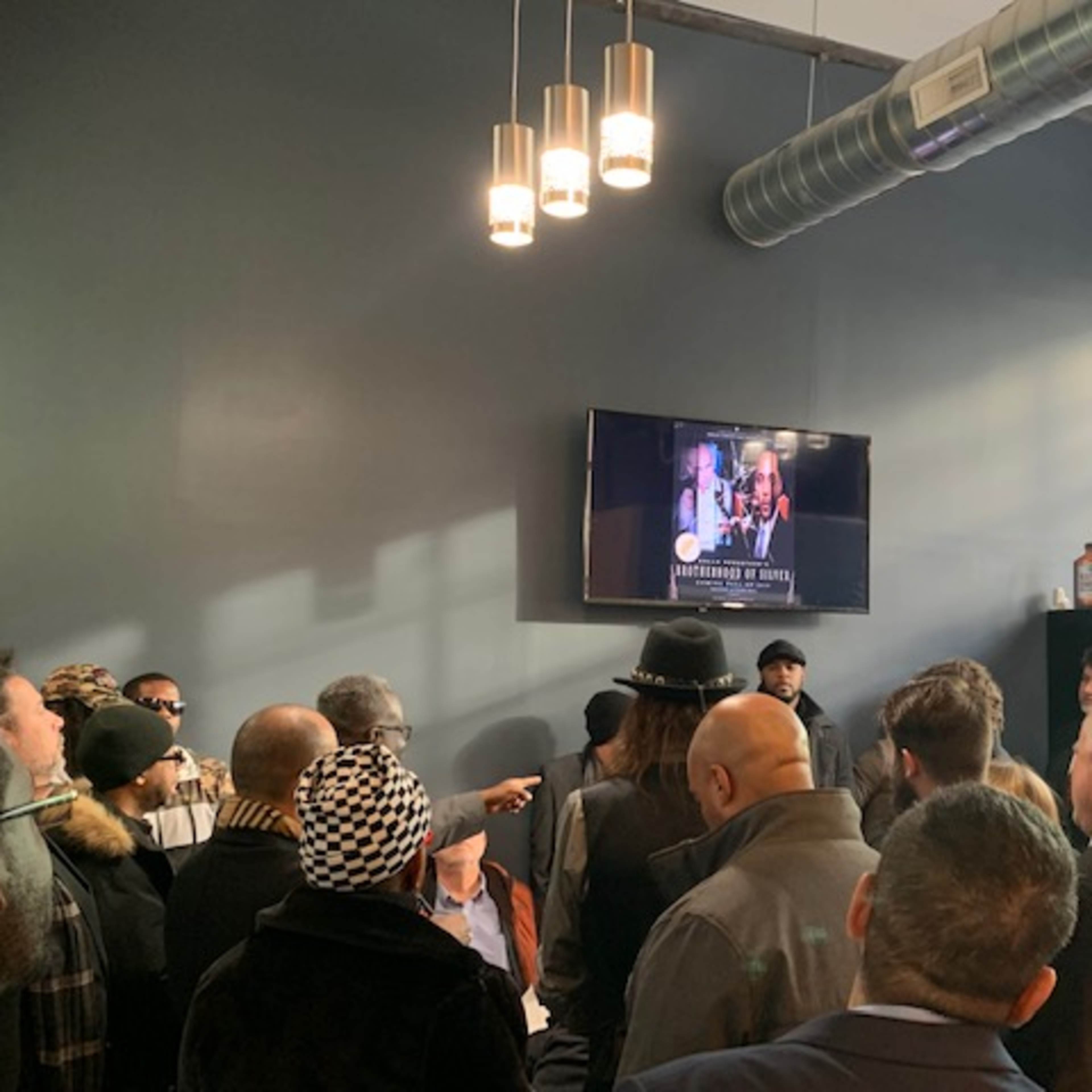 A crowd of people gathers indoors, watching a video displayed on a wall-mounted television screen.